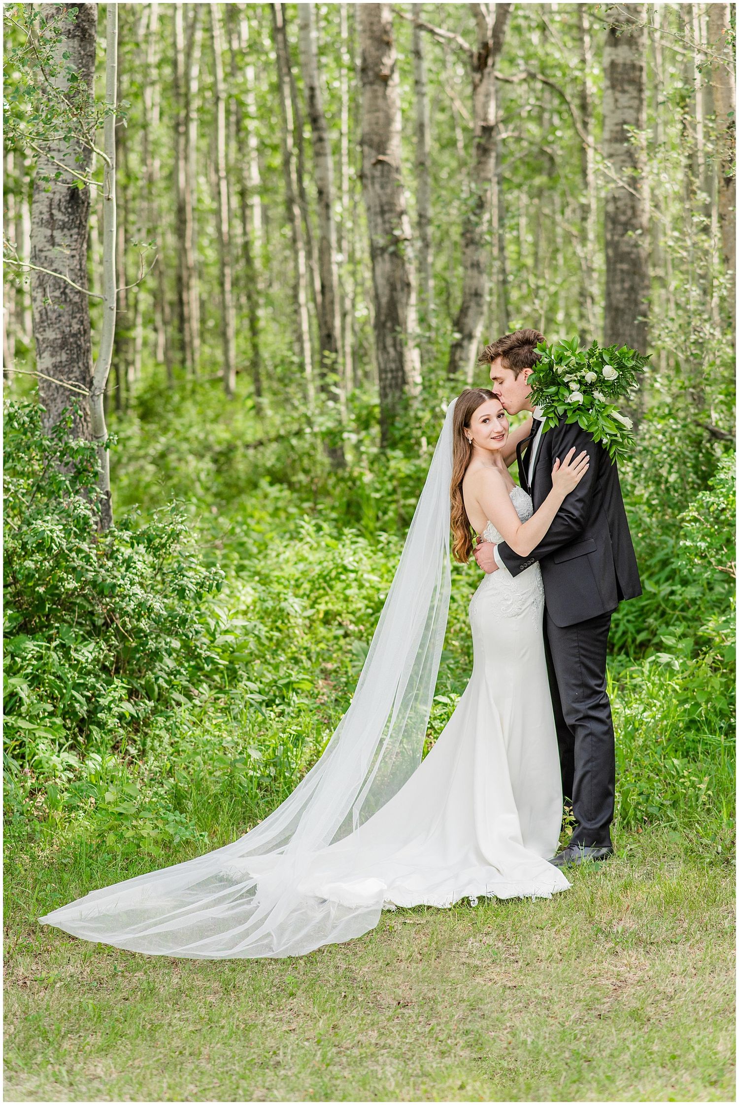 Bride and groom embrace in lush green forest, her long veil trailing on grass.