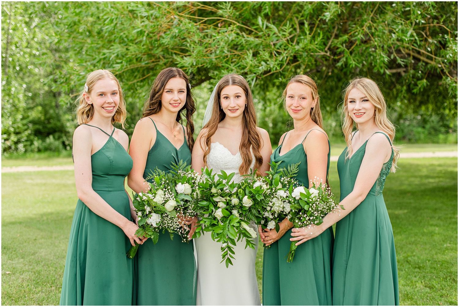 Four bridesmaids in emerald green dresses flank the bride, holding white and greenery bouquets outdoors.