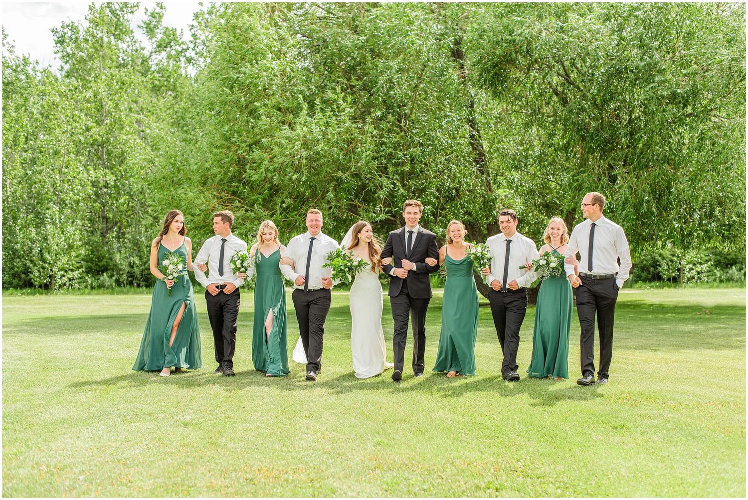Wedding party walks together in green bridesmaid dresses and black suits on a lush garden lawn.