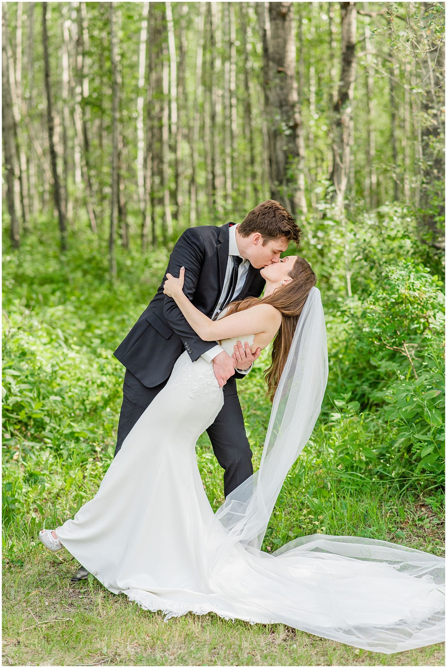 Groom dips bride for a romantic kiss in a lush green forest, her long veil trailing on the ground.