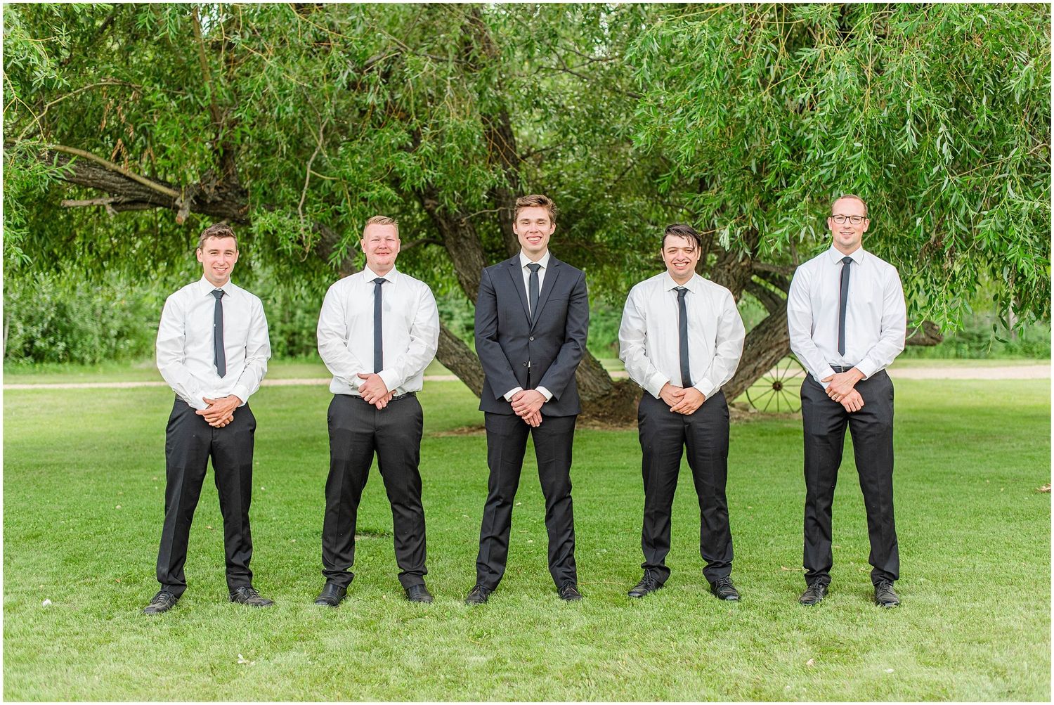 Groom stands with four groomsmen in black pants, white shirts, and dark ties outdoors.