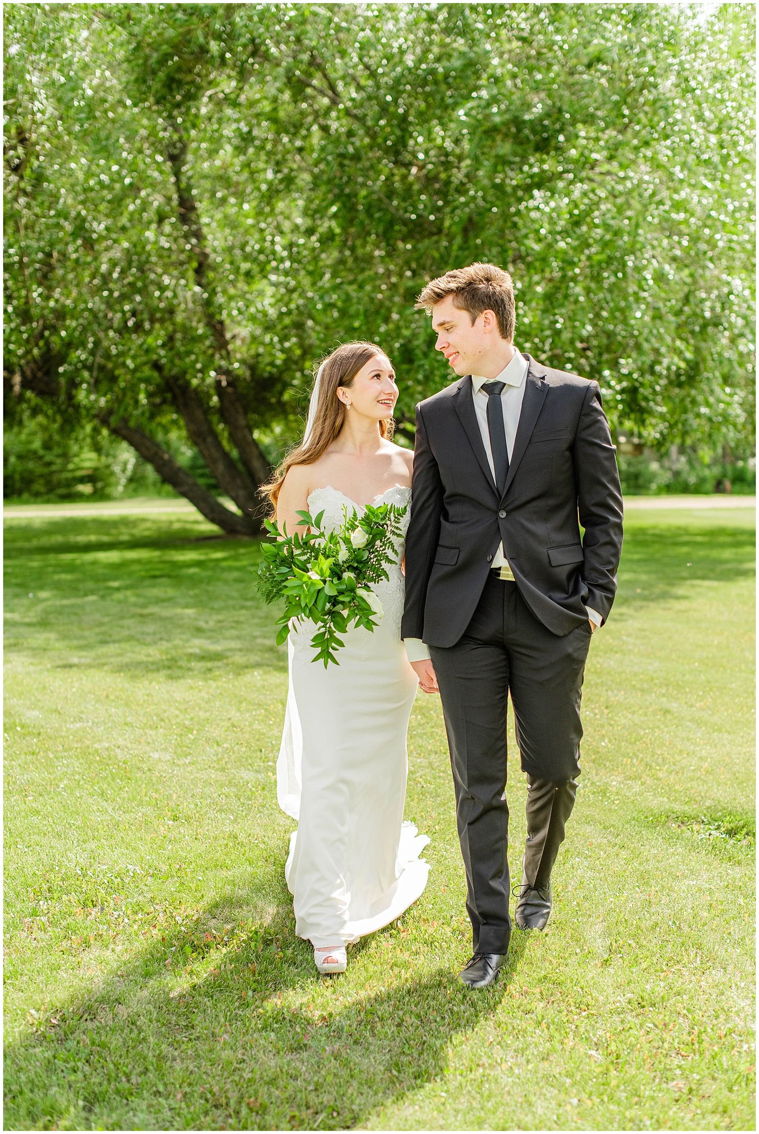 Bride in white gown holding green bouquet walks with groom in black suit on sunny lawn.