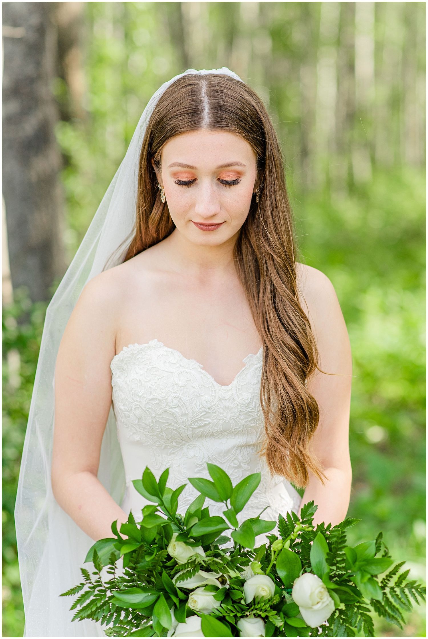 Bride in strapless lace gown holds lush green and white bouquet in woodland setting.