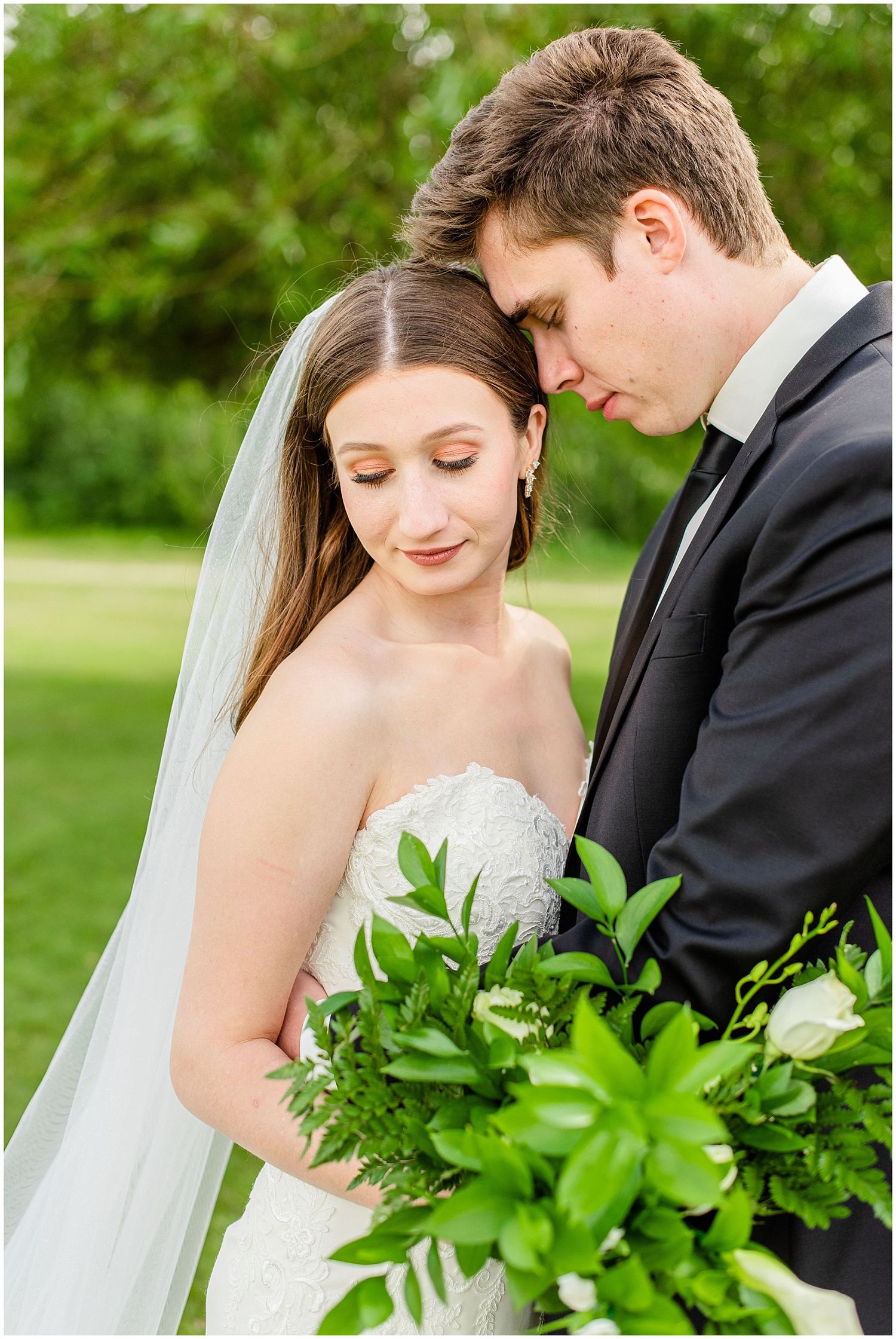 Bride and groom share an intimate moment, foreheads touching, as she holds a lush green bouquet.
