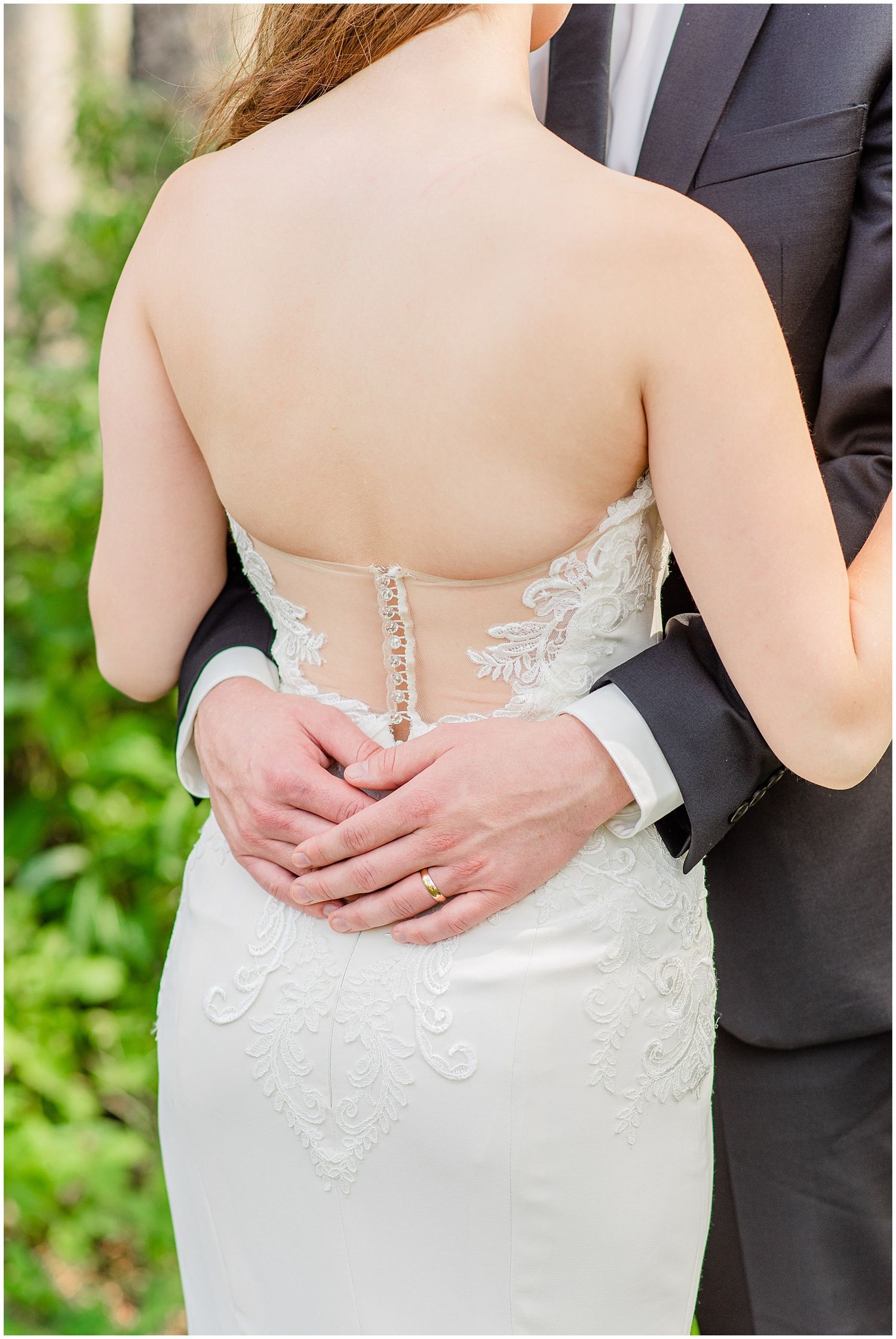 Close-up of groom's hands around bride's waist, showcasing intricate lace back detail of wedding gown.