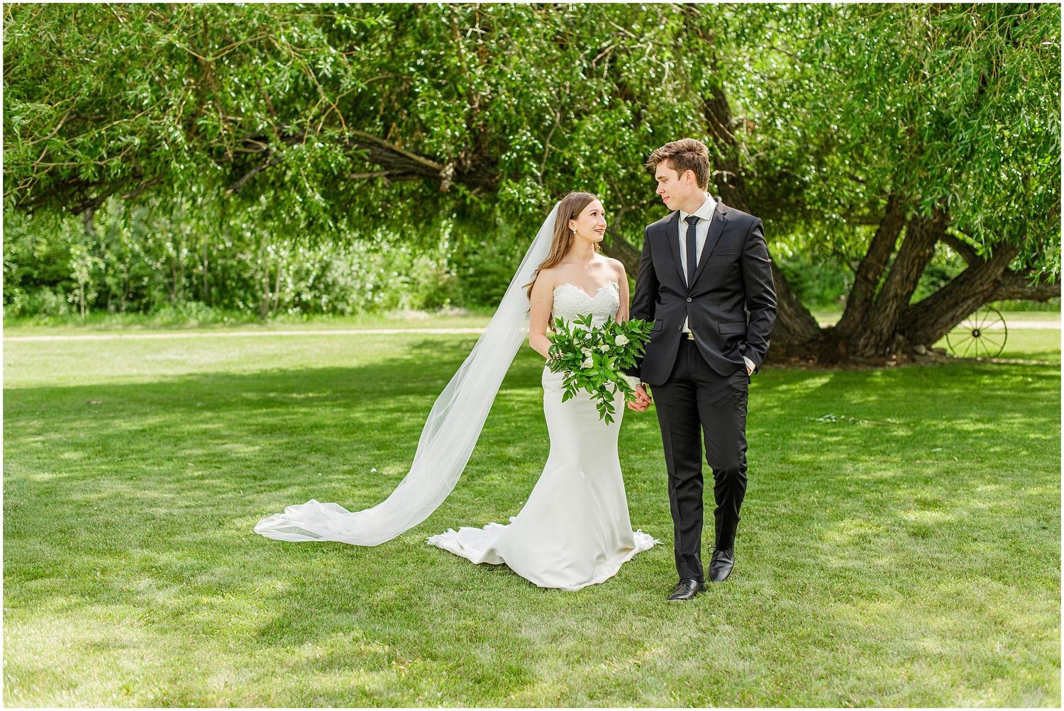Bride and groom walk hand in hand across a green lawn, bride wearing a long veil trailing behind her.