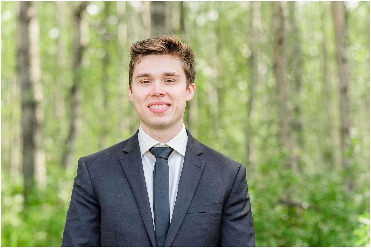 Young man in a dark suit and tie smiling in front of birch trees at a wedding.