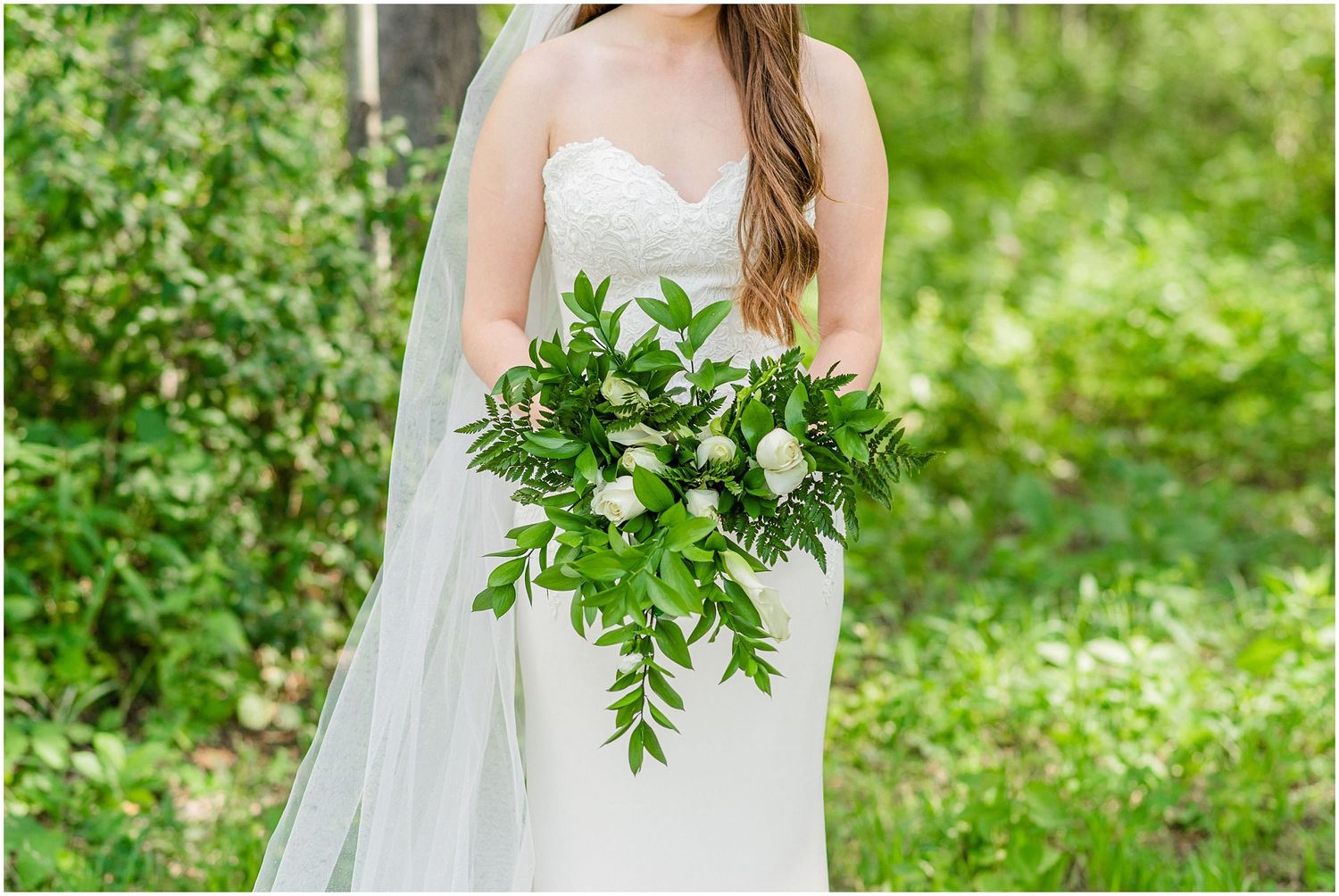 Bride in strapless white gown holds a lush bouquet of white roses and cascading greenery in a garden.