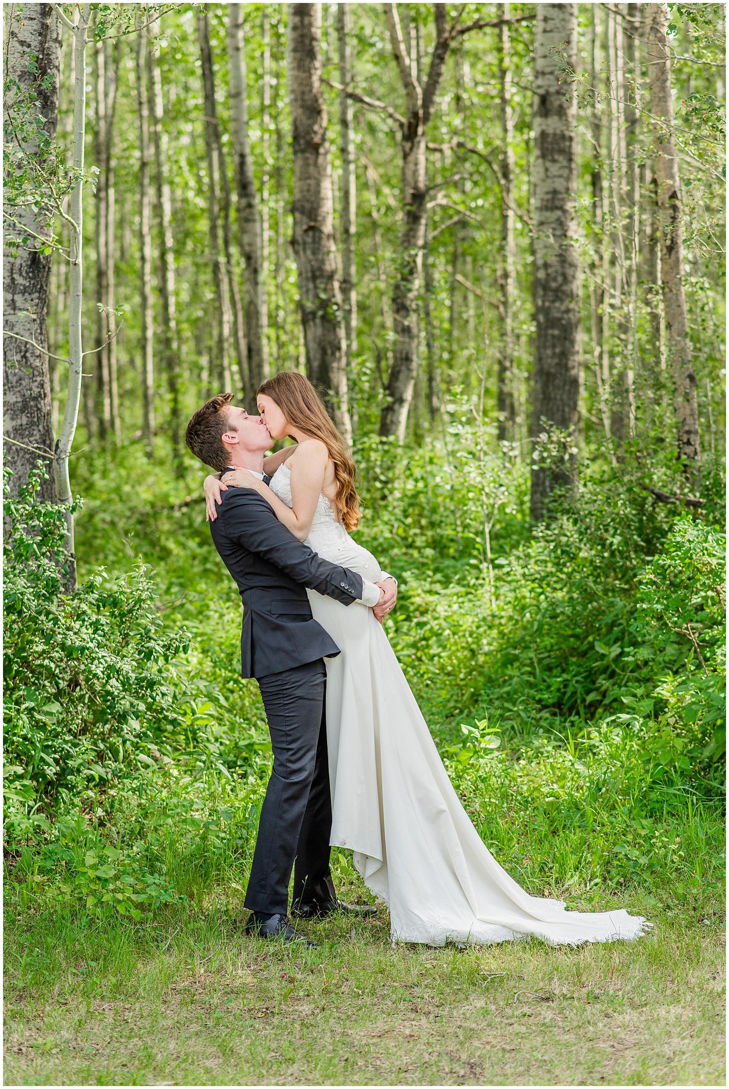 Groom lifting and kissing his bride in a white dress among lush green birch trees.