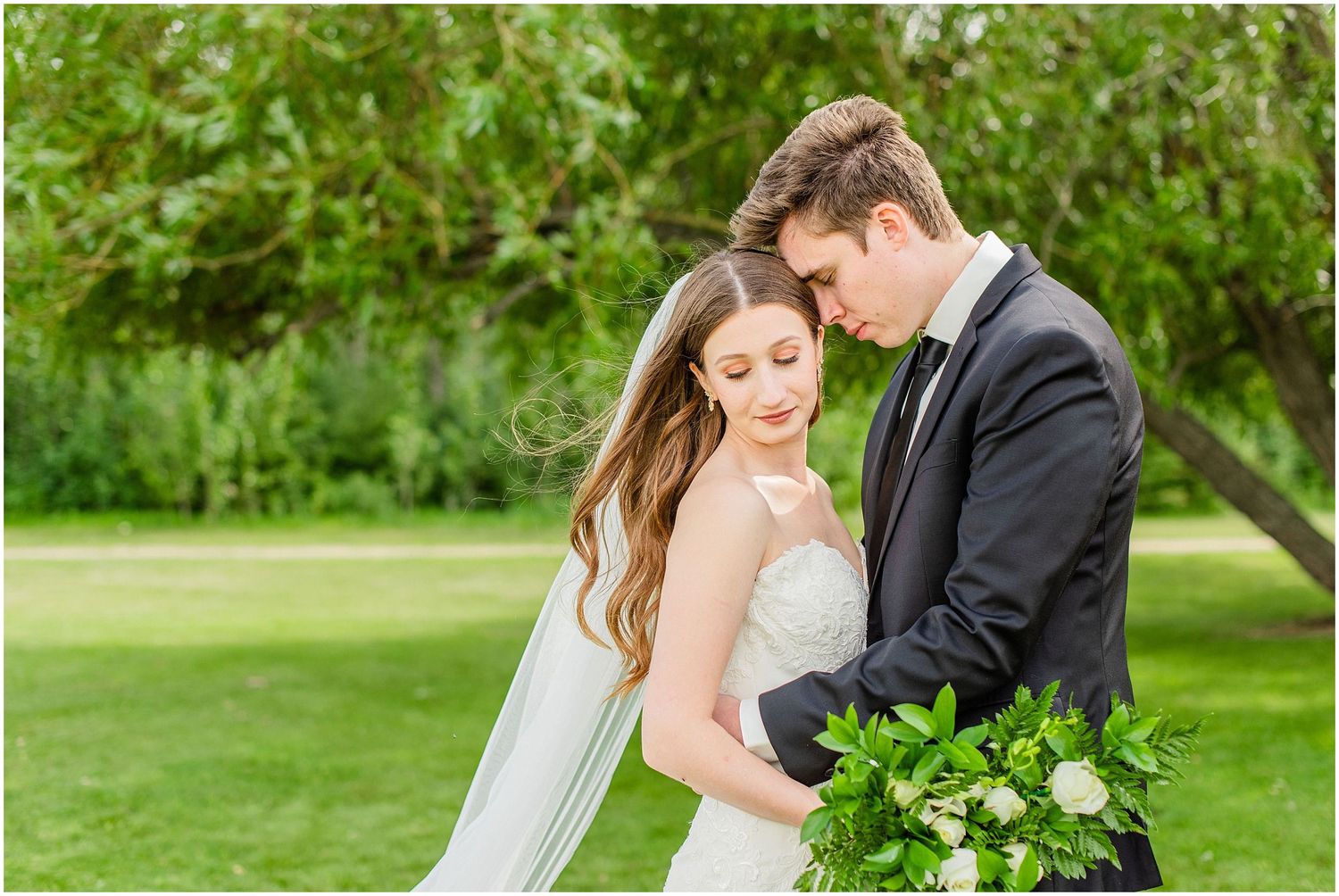 Bride and groom share an intimate moment, foreheads touching, as he holds her lush green and white bouquet.