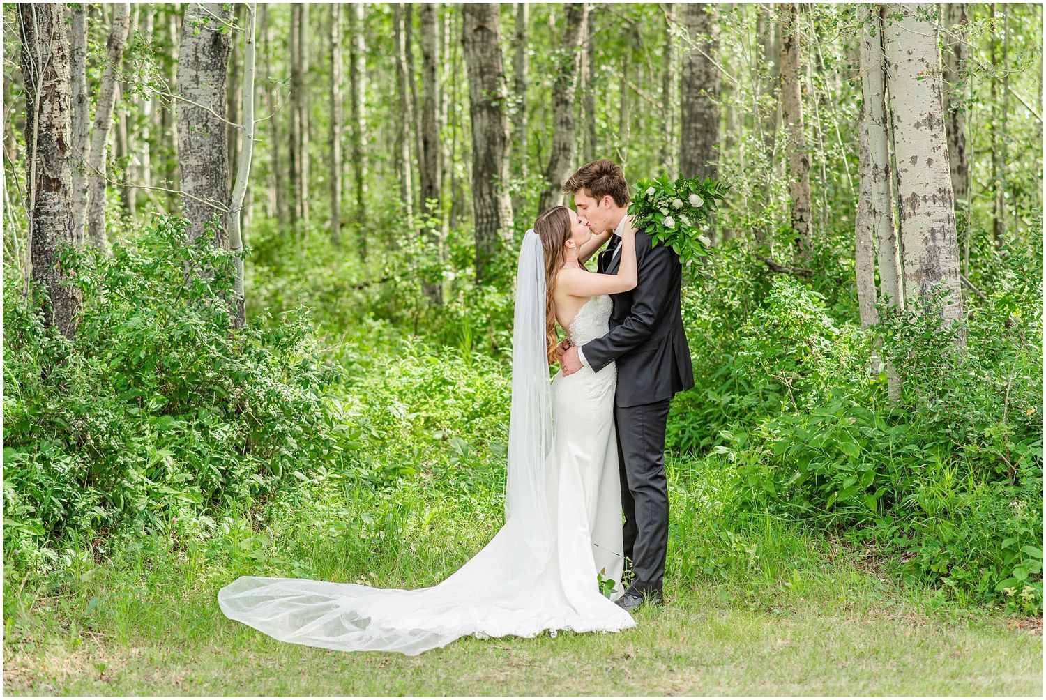Bride and groom embrace romantically in a lush green birch forest setting.