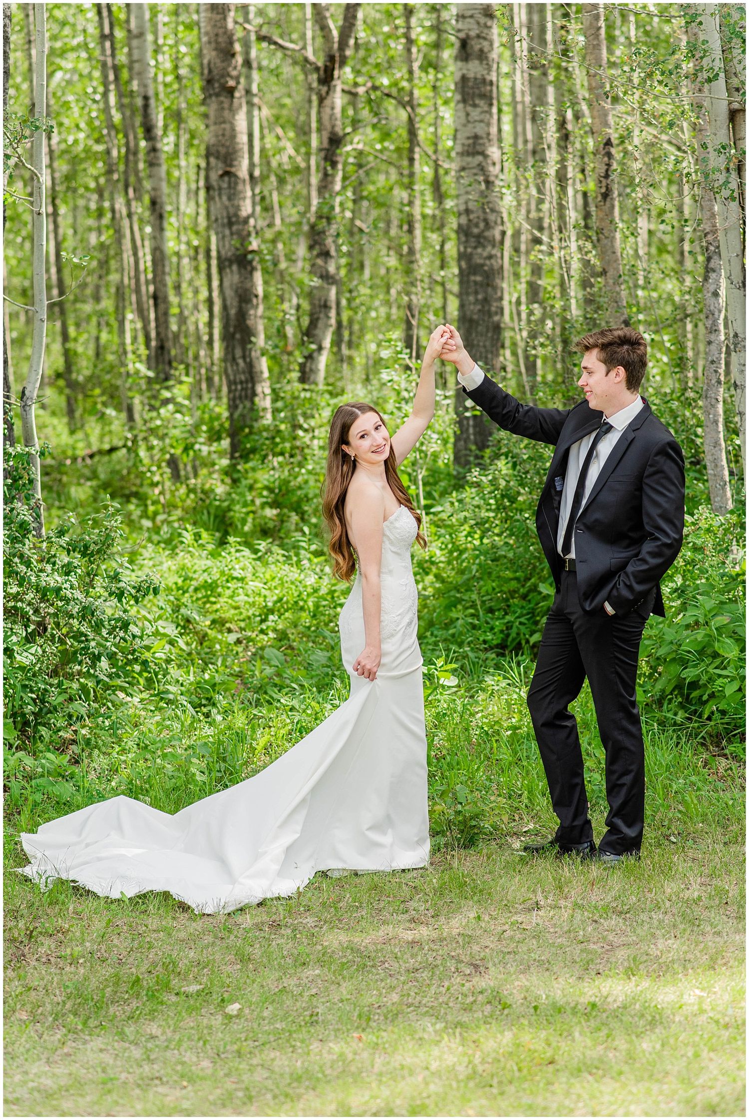 Bride and groom dancing among birch trees, bride in elegant white gown with train, groom in black suit.