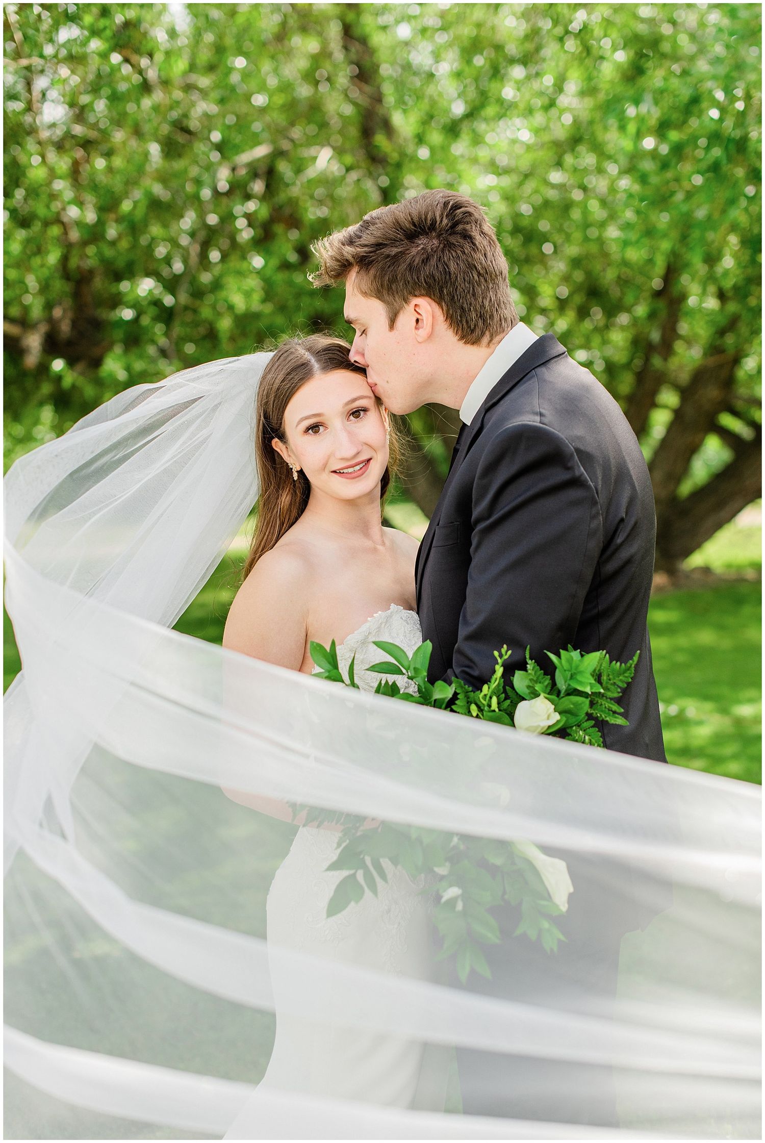 Groom kissing bride on the cheek as her long veil dramatically billows around them outdoors.