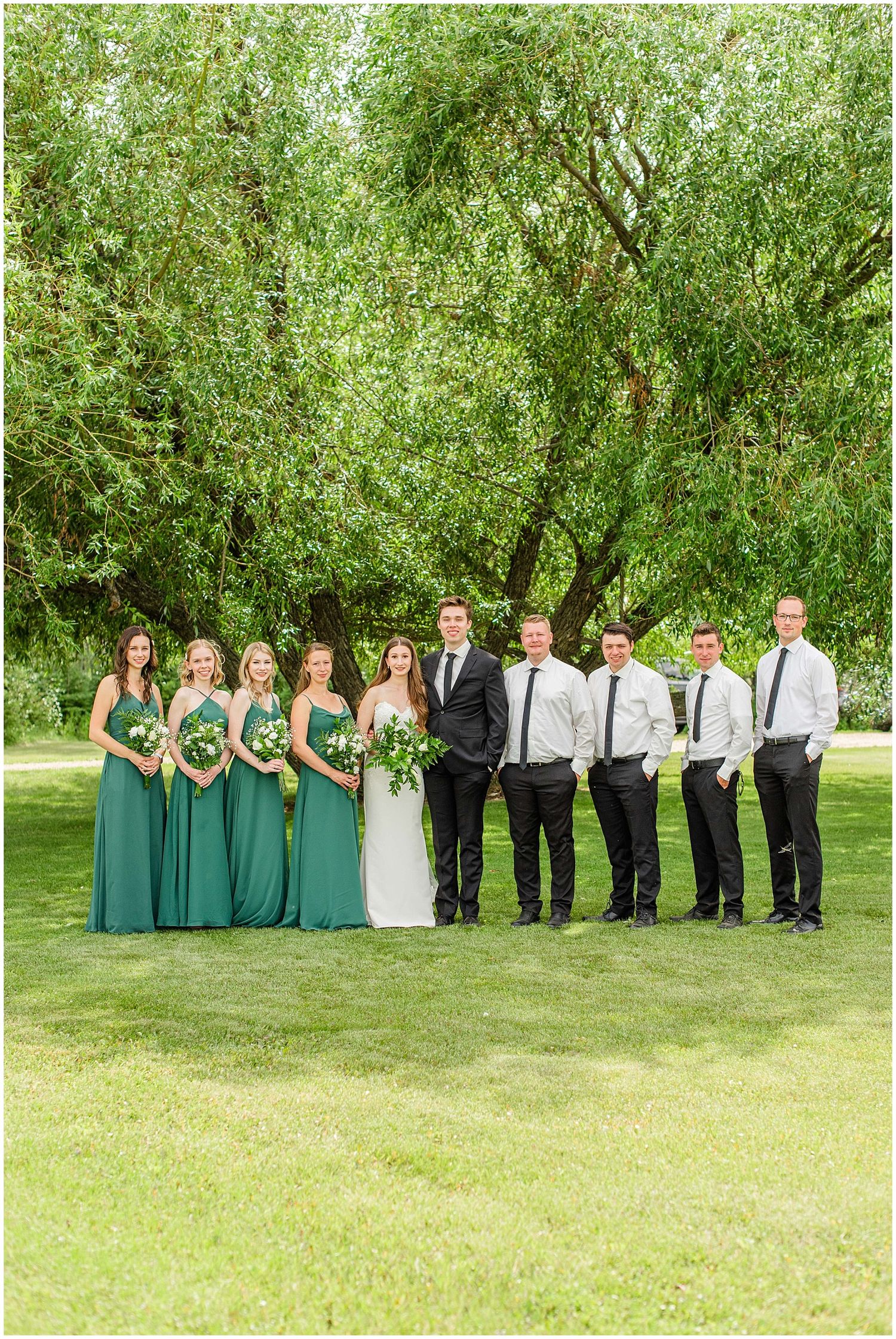 Wedding party poses outdoors with bridesmaids in emerald green gowns and groomsmen in black.