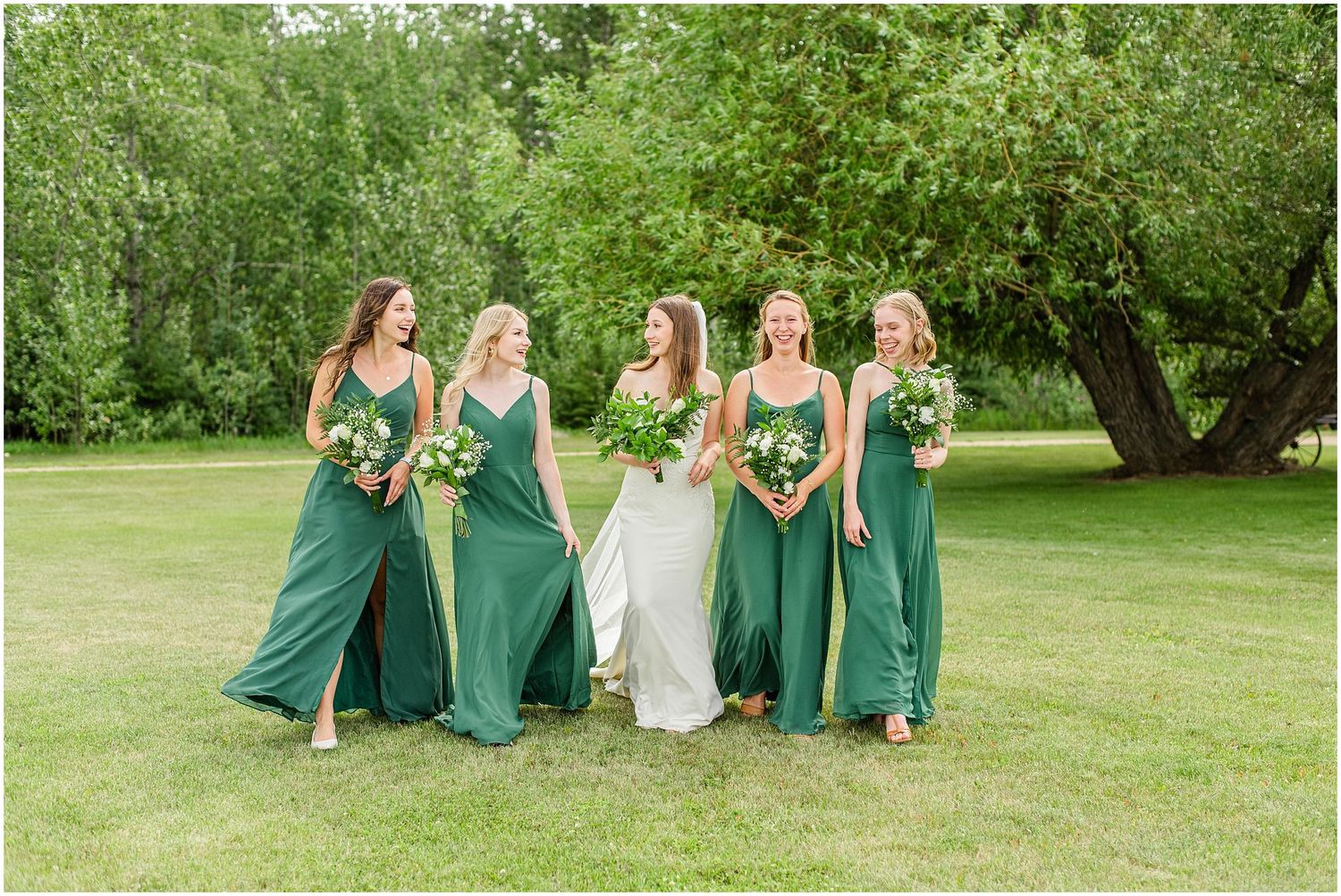 Bride and four bridesmaids in flowing emerald green dresses walk together laughing in a sunlit garden.