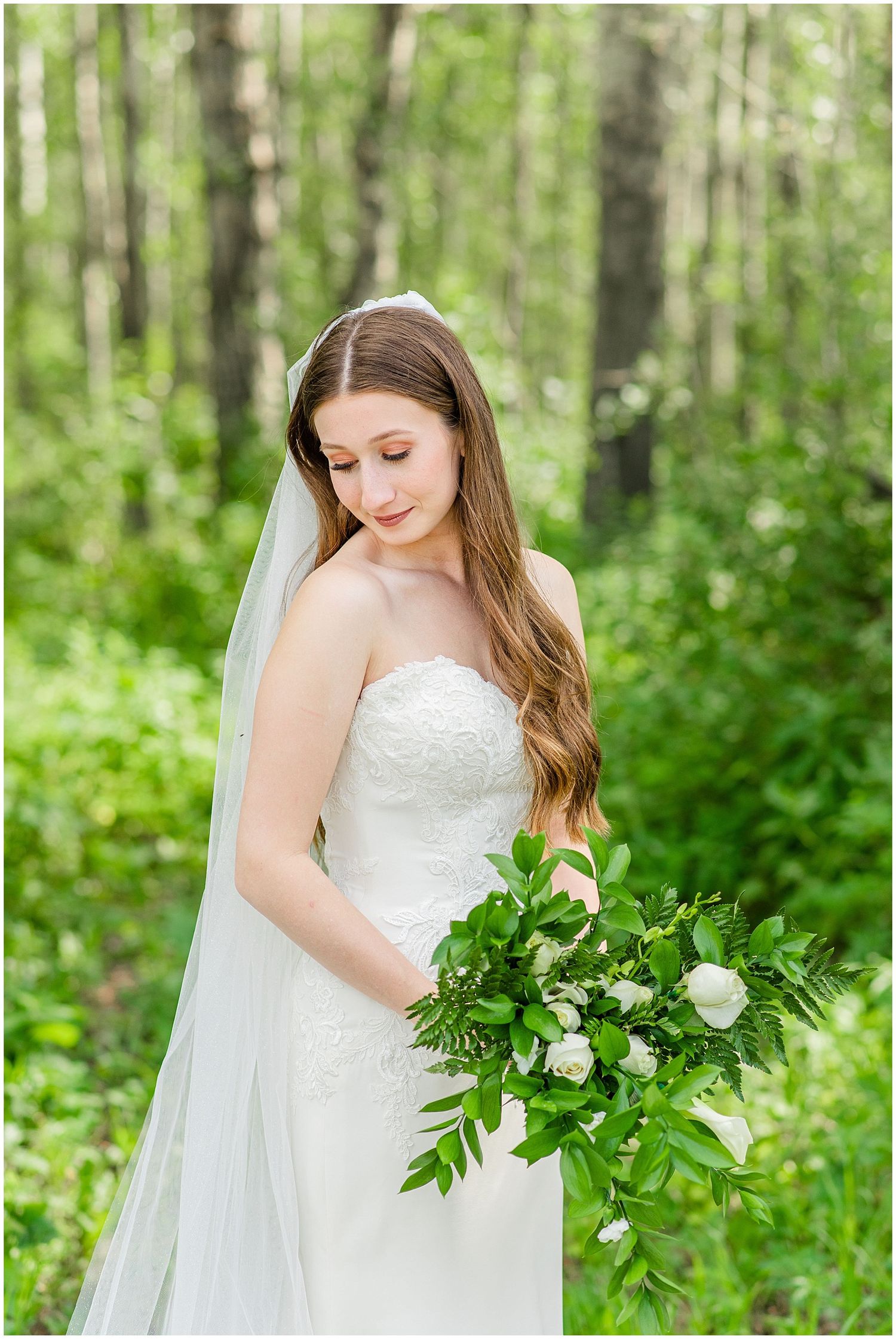 Bride in a strapless lace gown holds a green and white bouquet while standing in a lush forest.