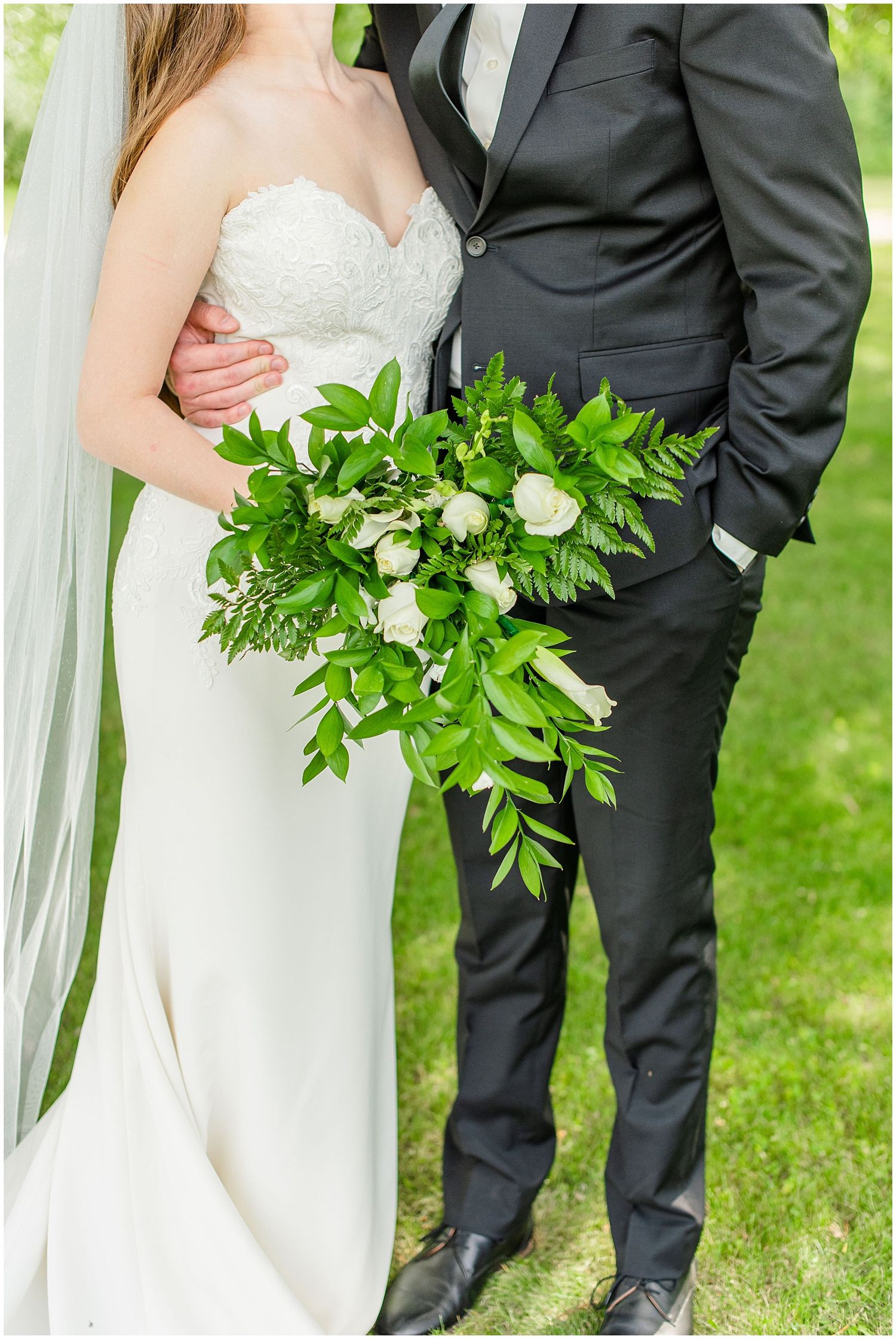 Close-up of bride in lace gown holding lush green and white bouquet with groom.