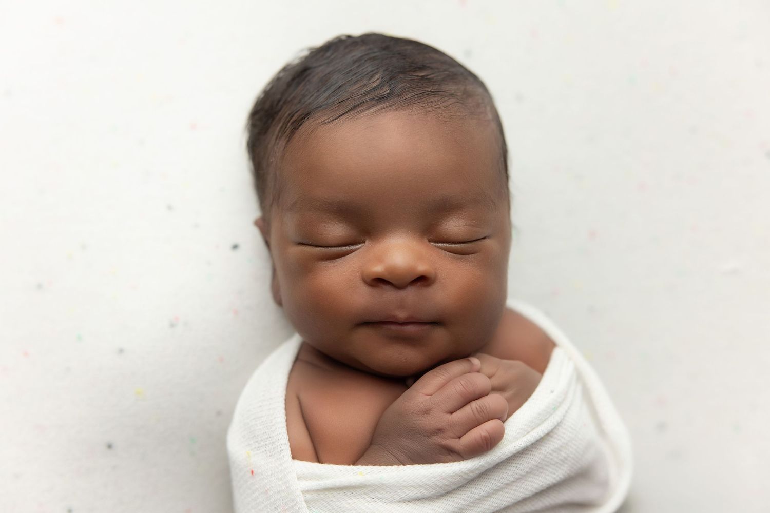 Peaceful newborn baby swaddled in white wrap, sleeping with eyes closed on a light background.