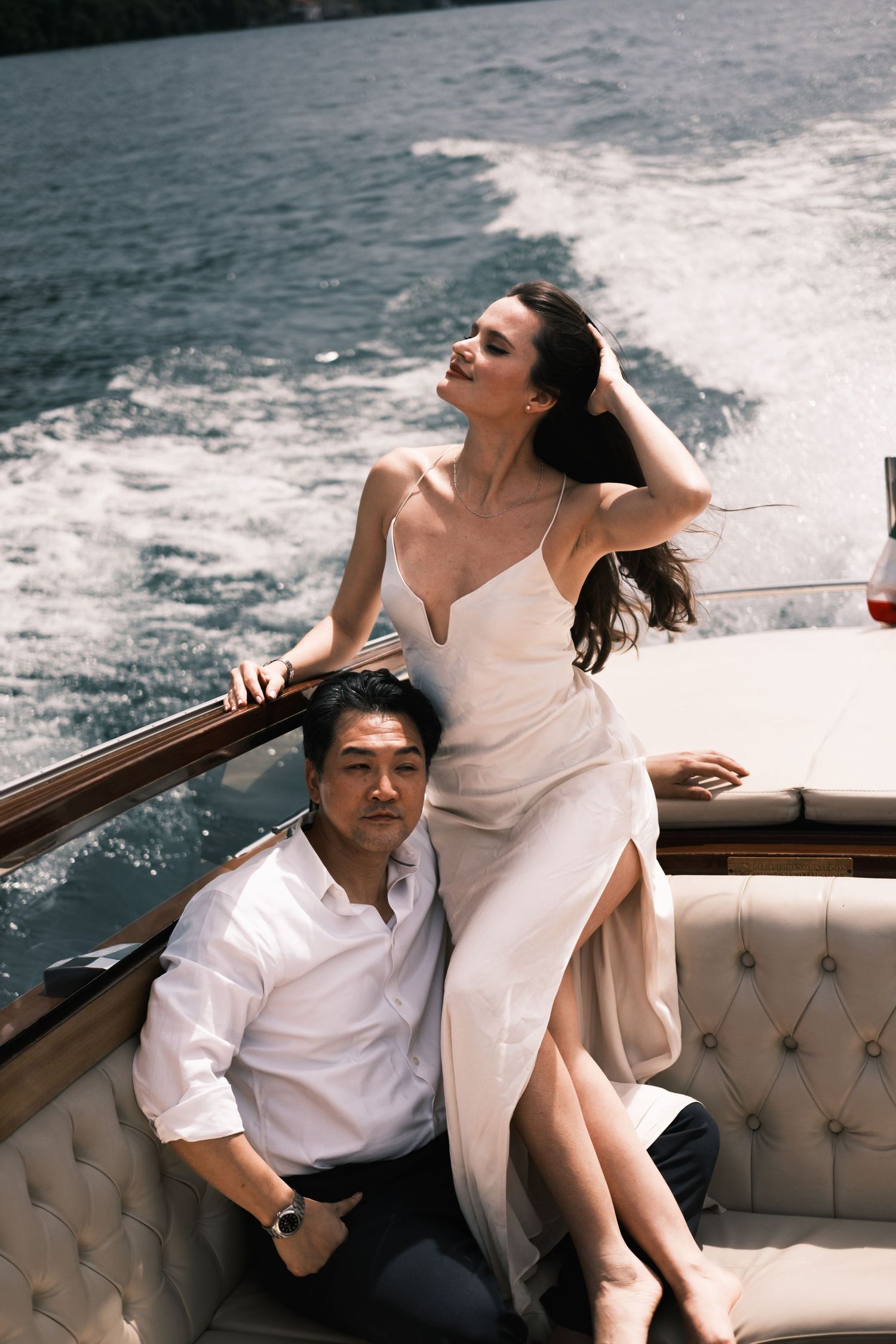 Elegant couple on a luxury speedboat, woman in white slip dress posing, man in white shirt seated, ocean backdrop.