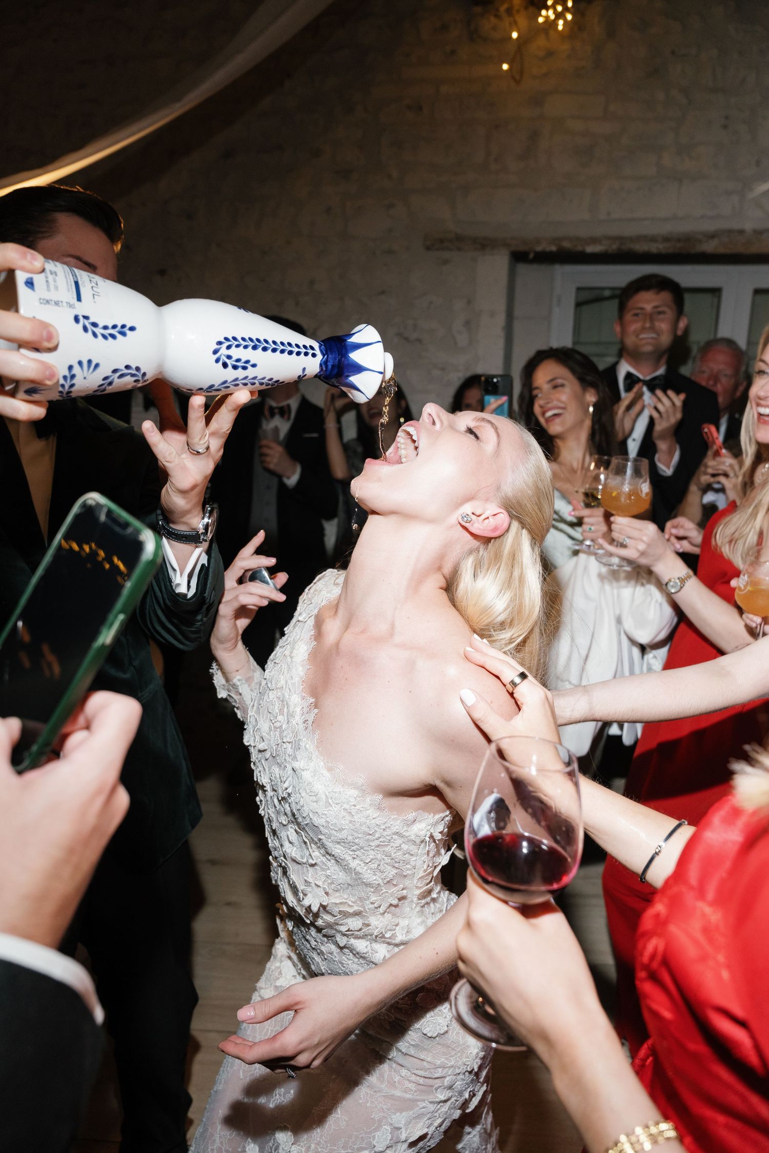Bride in lace gown drinking tequila from bottle at lively wedding reception celebration.