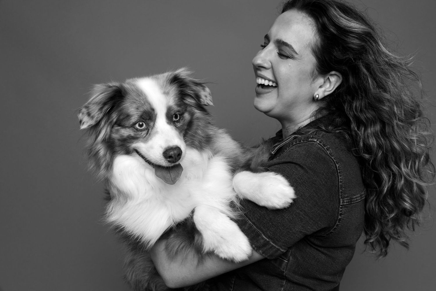Black and white photo of a smiling woman holding a red merle Australian Shepherd dog against a gray background.