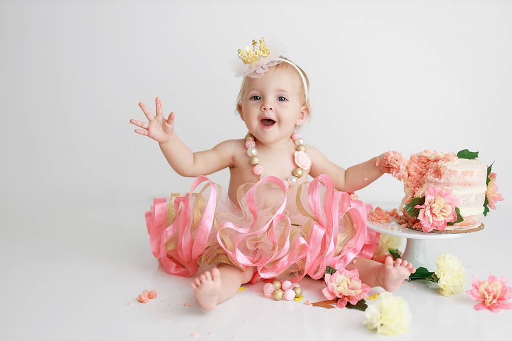 Happy baby girl in pink ribbon tutu and pearl necklace celebrates first birthday with floral cake by Winchester baby photographer.