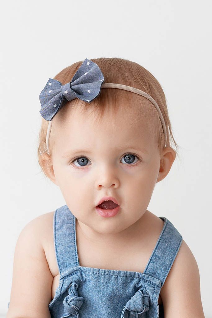 Adorable baby girl with blue eyes wearing denim overalls and polka dot bow headband on white background.