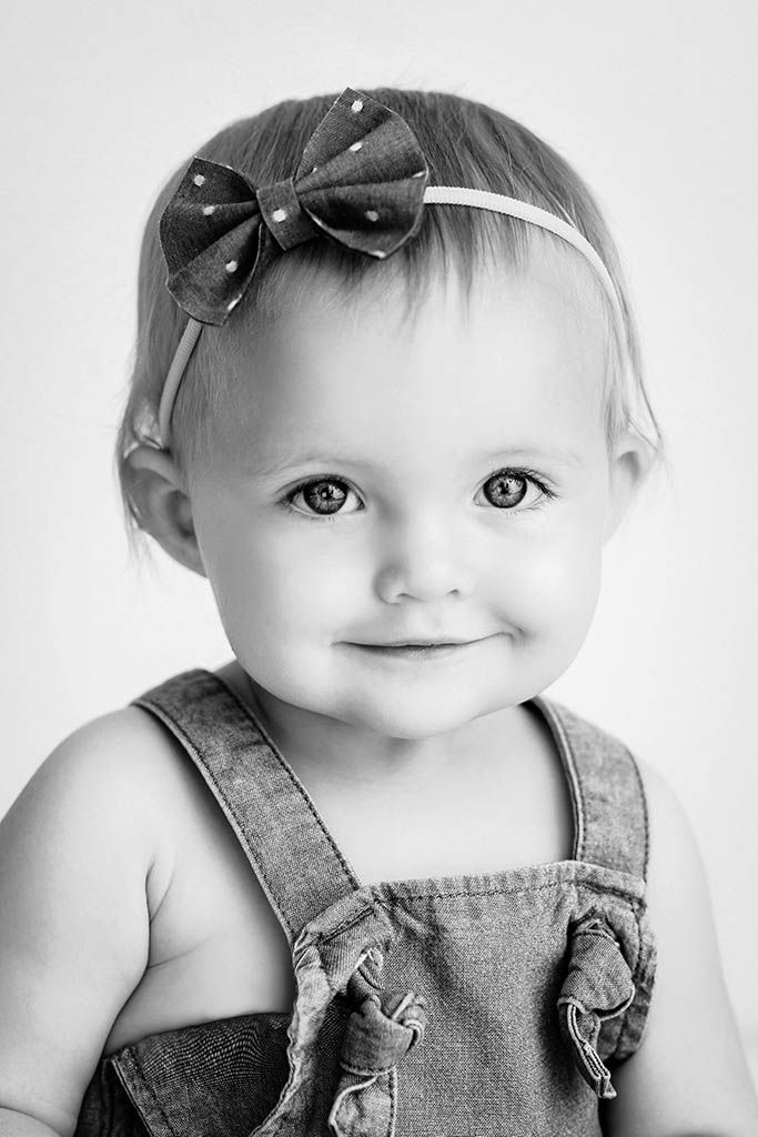 Winchester baby photography - Black and white portrait of a smiling baby girl wearing a bow headband and denim overalls.