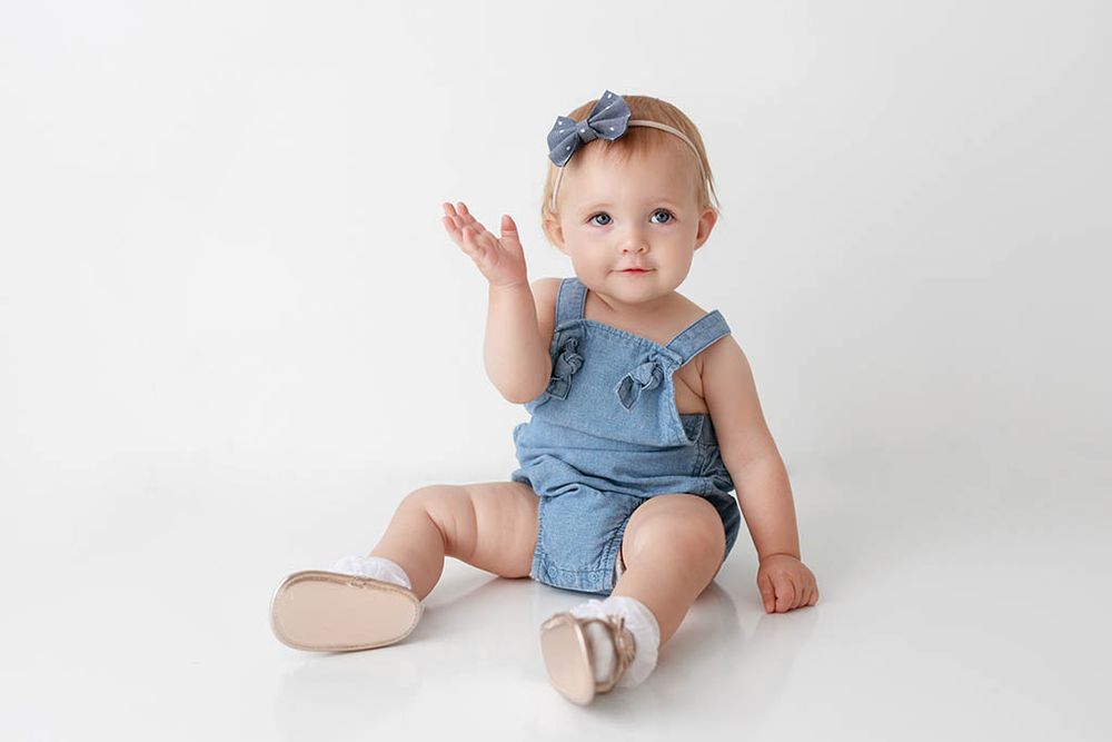 Baby girl in blue denim romper and bow headband sitting on white background, waving her hand.