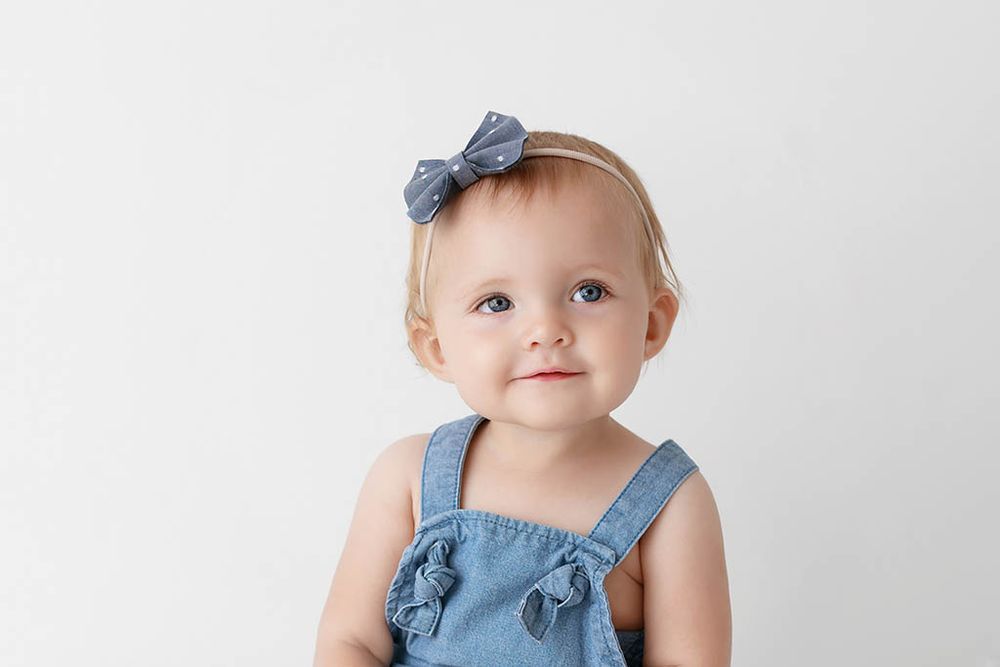 Adorable baby girl portrait in denim romper with blue bow headband smiling against white backdrop by Winchester baby photographer.