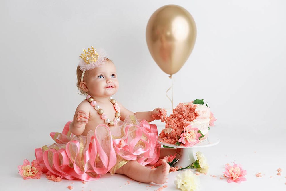 One year old baby girl in pink tutu and crown sits with cake and balloon on white background by Winchester baby photographer.