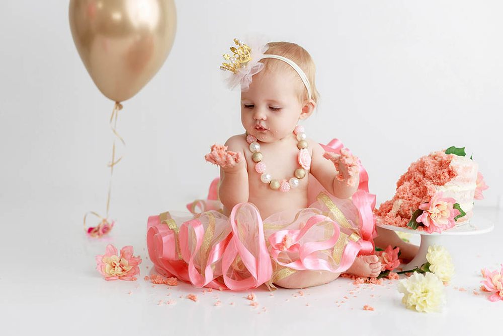 Baby girl in pink tutu with gold crown blowing kisses during cake smash with flowers and balloon.