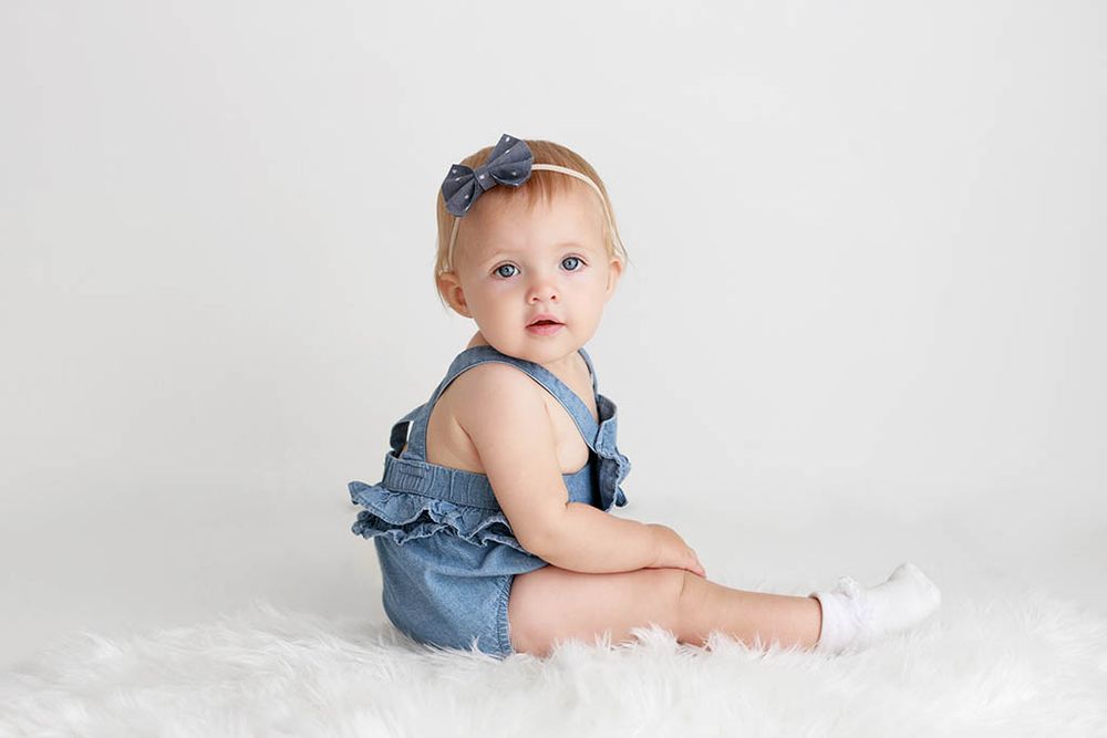 Adorable baby girl in denim ruffle romper and bow headband sits on white fur rug against light background by Winchester baby photographer.