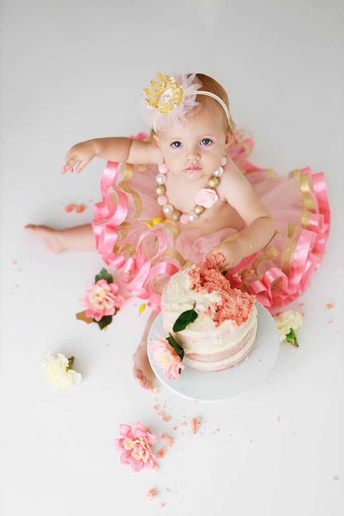 Baby girl in pink tutu and gold crown sits beside smashed first birthday cake with floral decorations.
