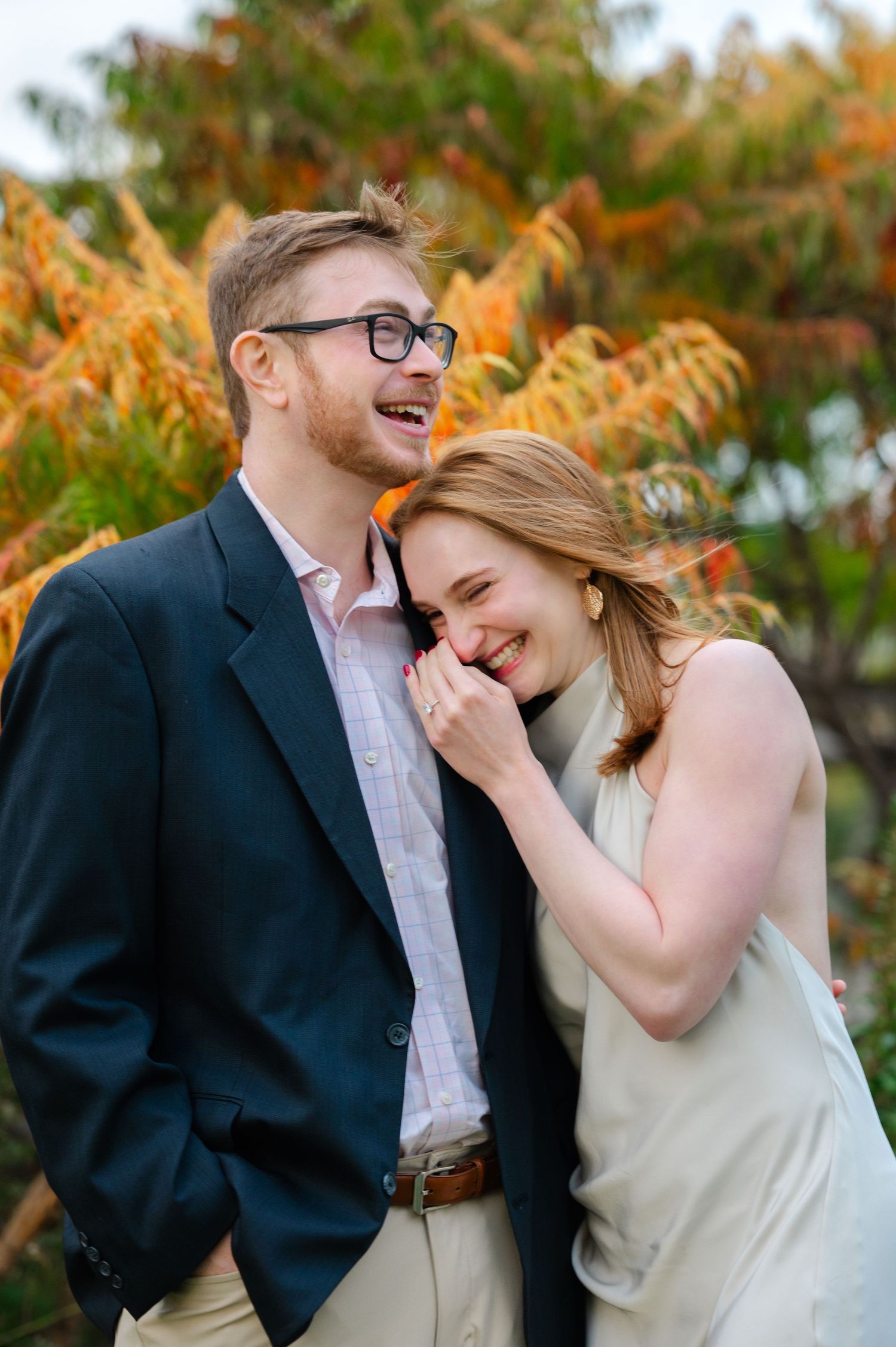 A laughing couple poses outdoors in front of autumn foliage, woman in a white dress leans on man's shoulder.