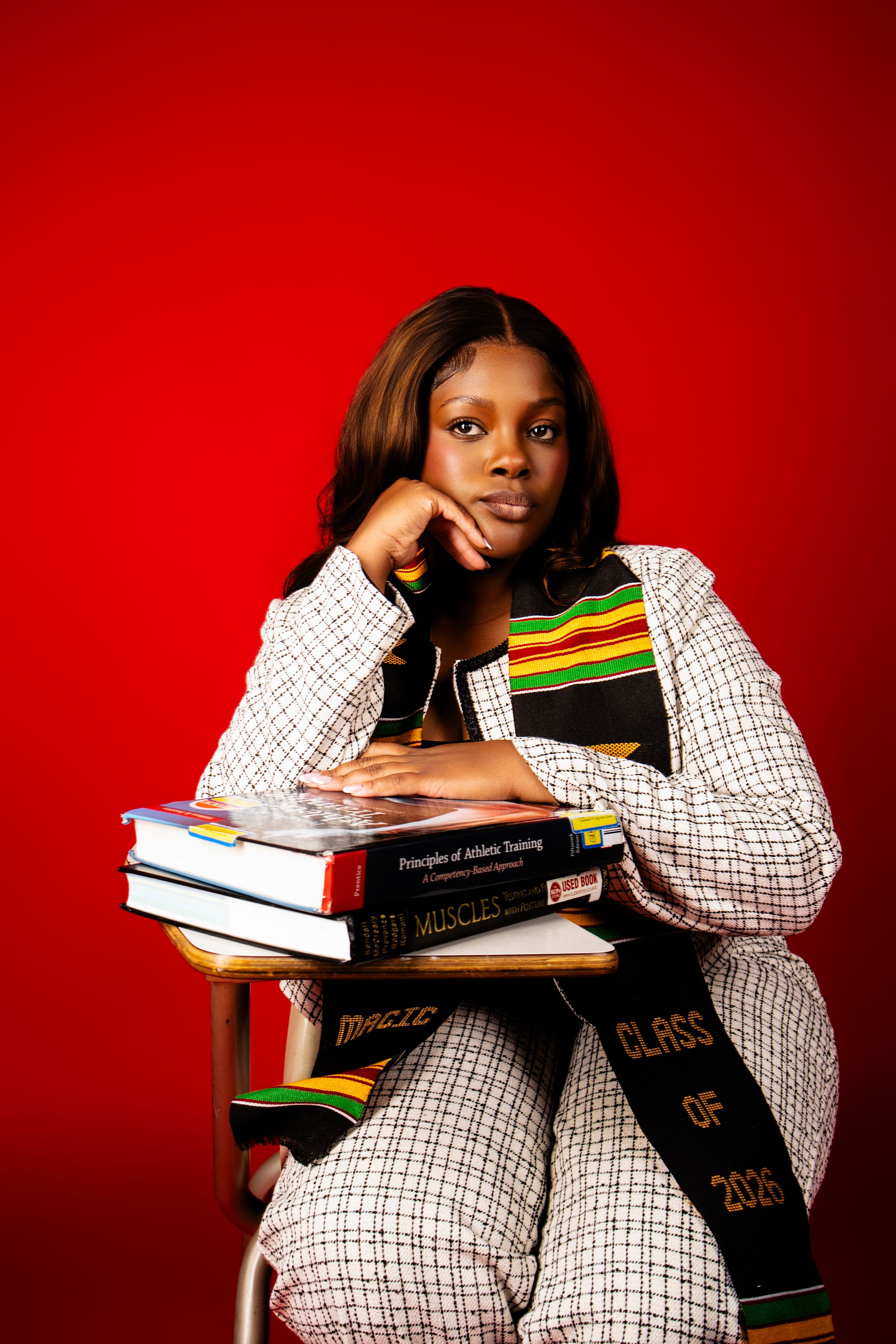Young woman in white blazer with graduation stole sits at desk holding books including Class of 2026 on red background.