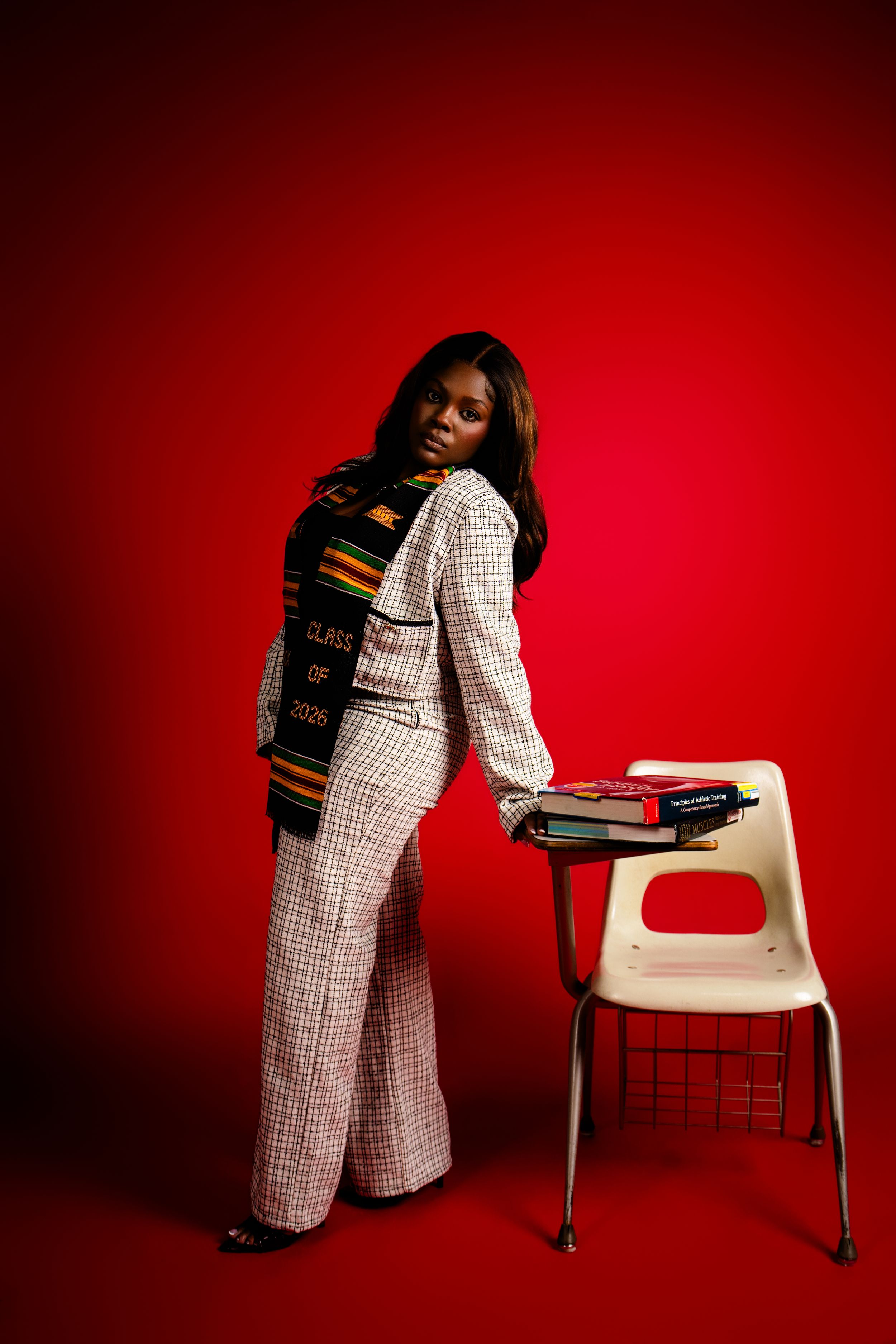 Graduate in plaid suit wearing Class of 2025 kente stole poses by school desk against red background.