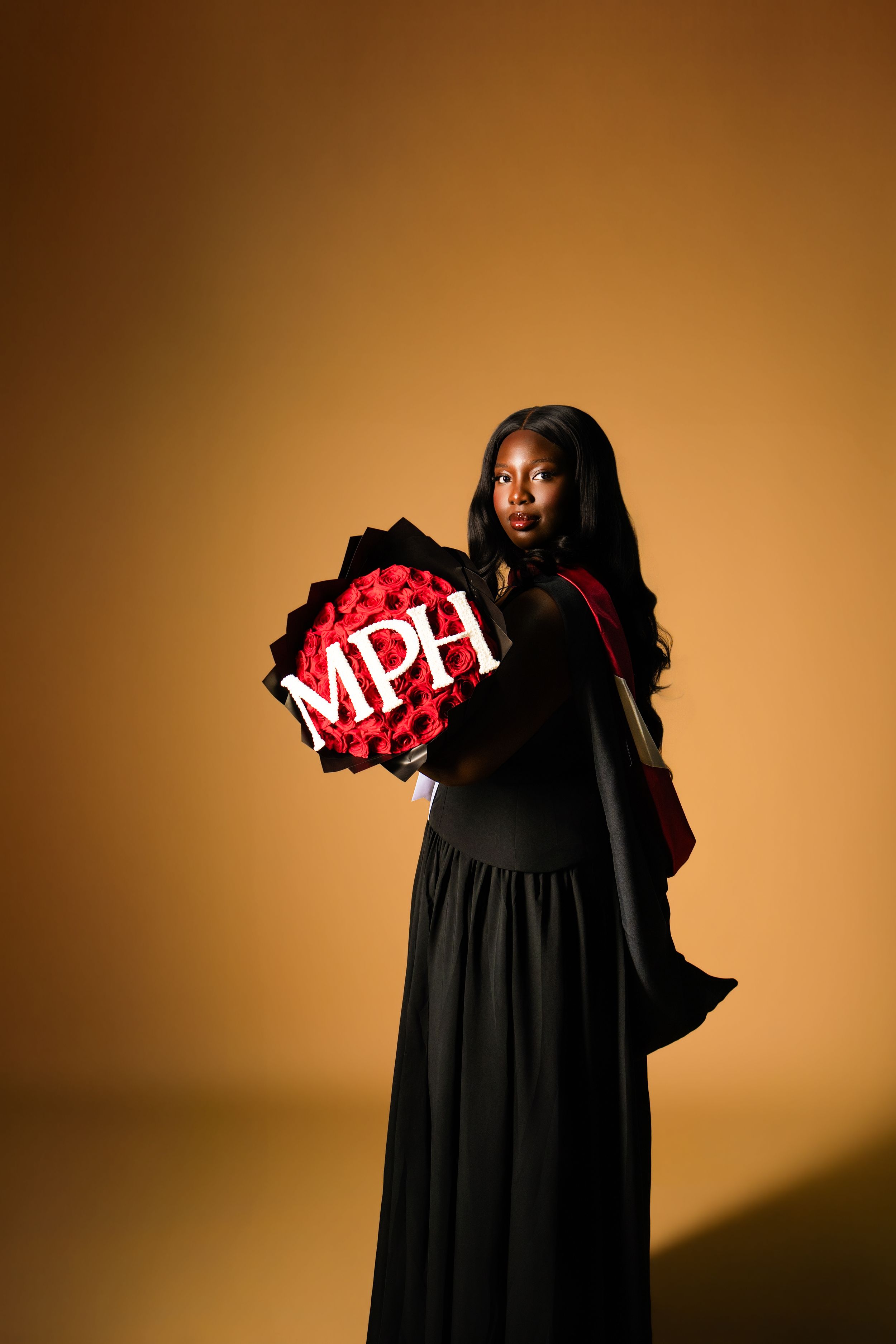 Woman in black graduation gown holding a decorative MPH sign with red roses on golden background.