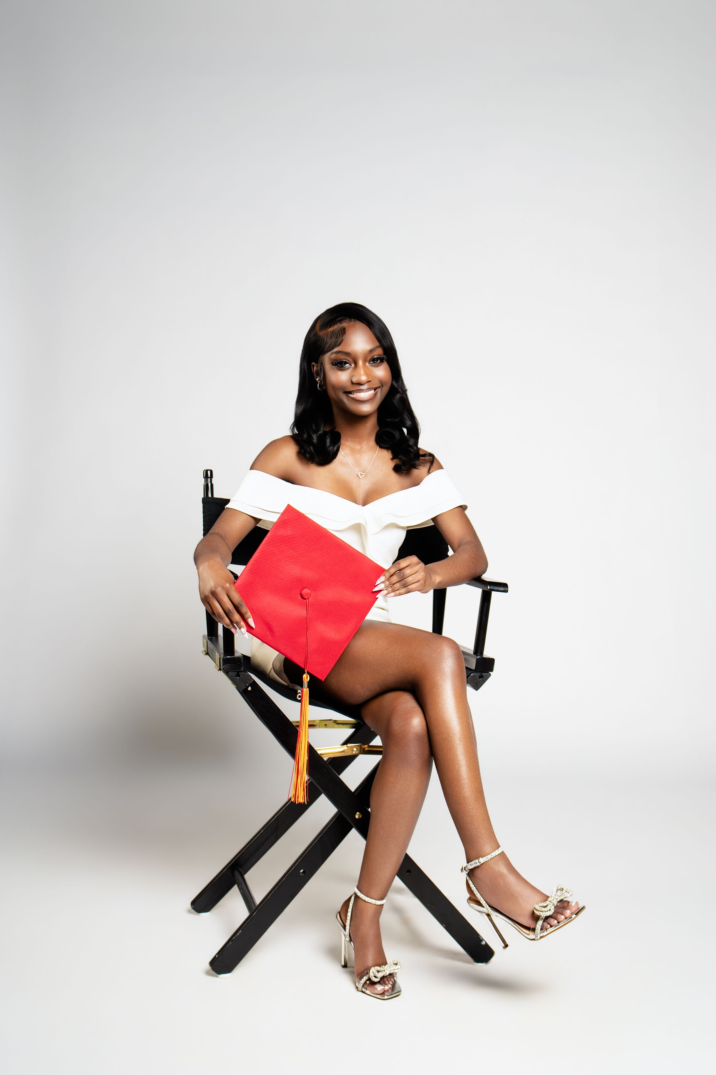 Smiling graduate in white dress holds red cap while seated in director's chair against white background.