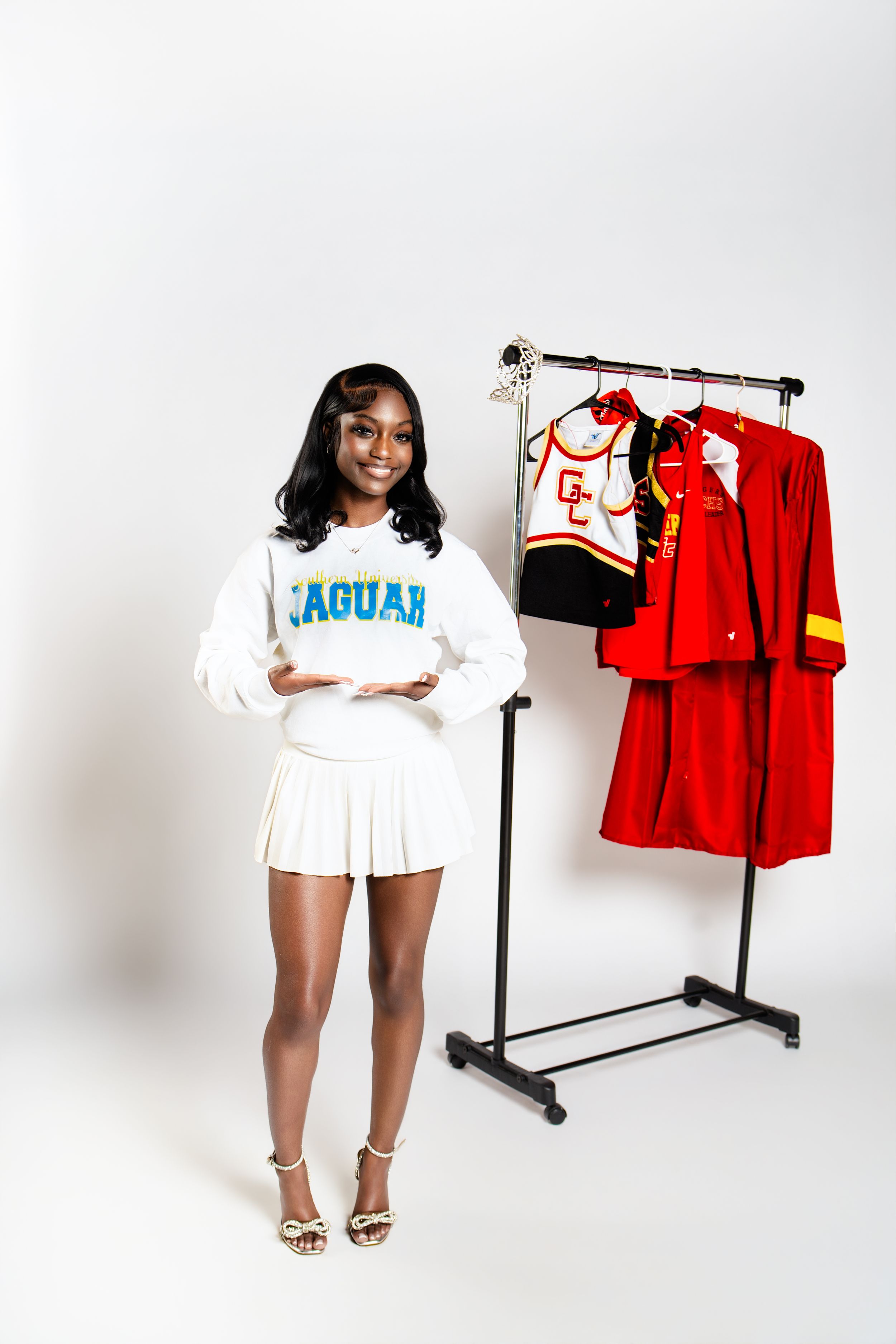 Young woman in Jaguar sweatshirt and white skirt poses next to clothing rack with red cheerleader uniforms.