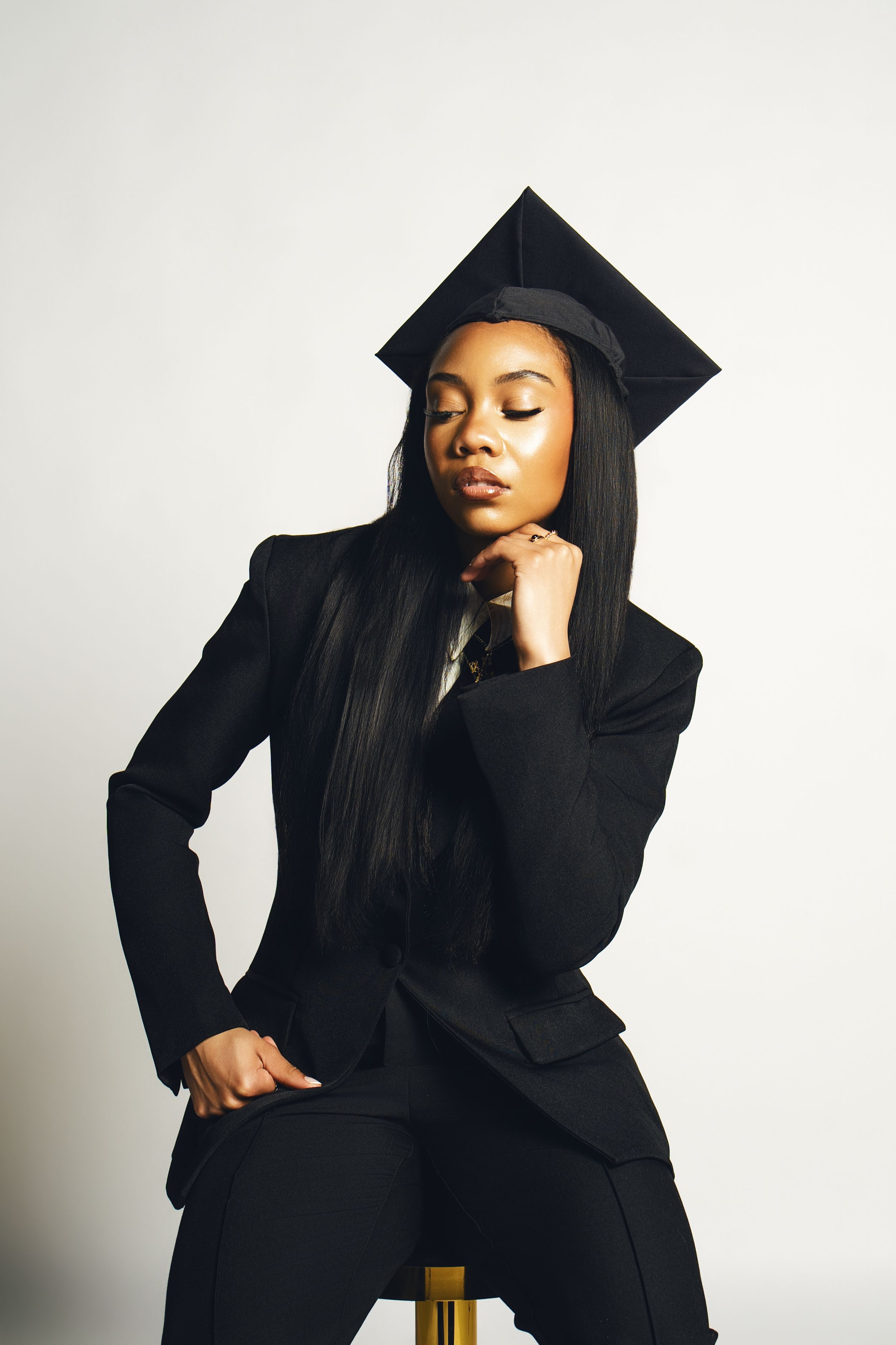 Young Black woman in black suit and graduation cap sits posed with eyes closed against white background.