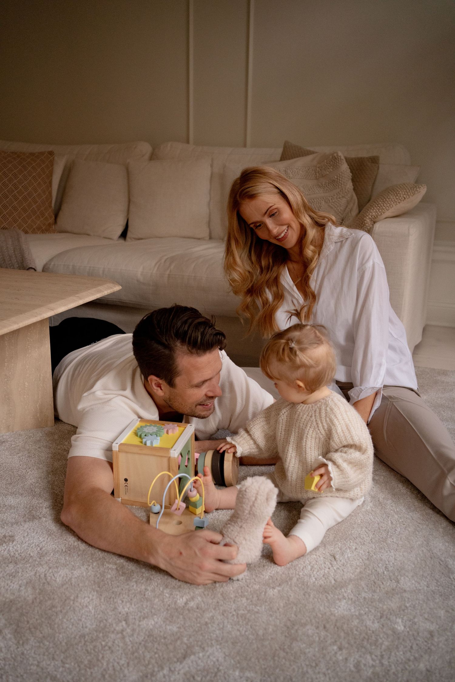 En mamma sitter och en pappa ligger på en matta framför en soffa och leker tillsammans med sitt barn som håller på en leksak.