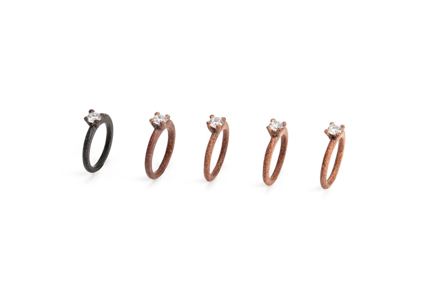 Set of five solitaire diamond rings ranging from black to rose gold on a white background.