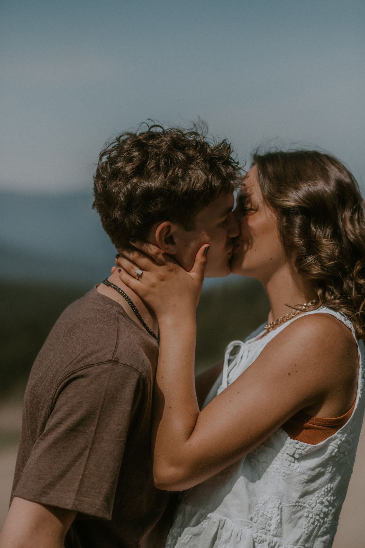 A young couple shares a romantic kiss outdoors with mountains in the background on a sunny day.