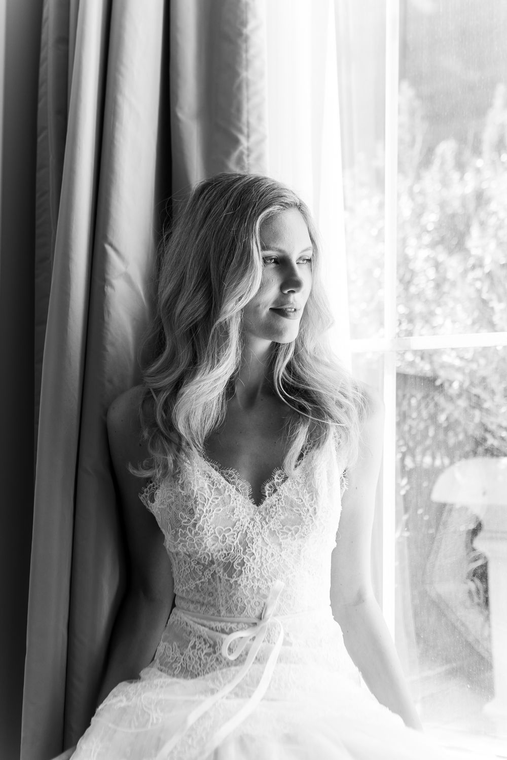Black and white portrait of a bride in lace wedding dress gazing out a window with soft natural light.