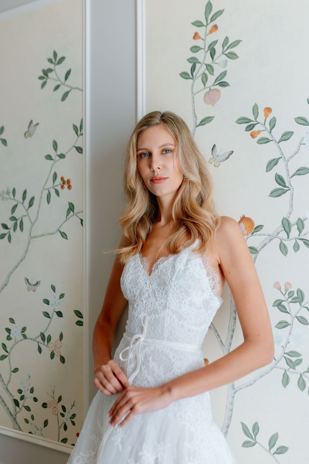 Blonde woman in white lace bridal gown with sweetheart neckline posing against floral wallpaper.