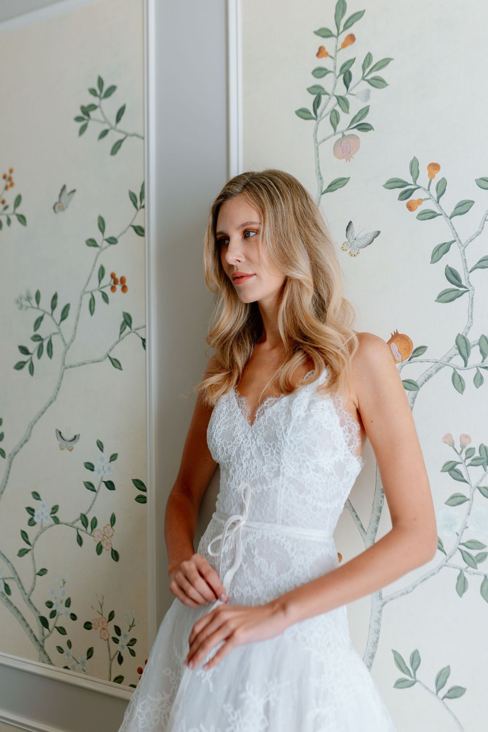 Elegant bride in white lace wedding dress with spaghetti straps standing by botanical wallpaper.