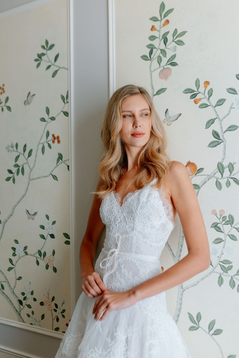 Blonde model in white lace bridal gown with spaghetti straps poses before floral chinoiserie wallpaper.