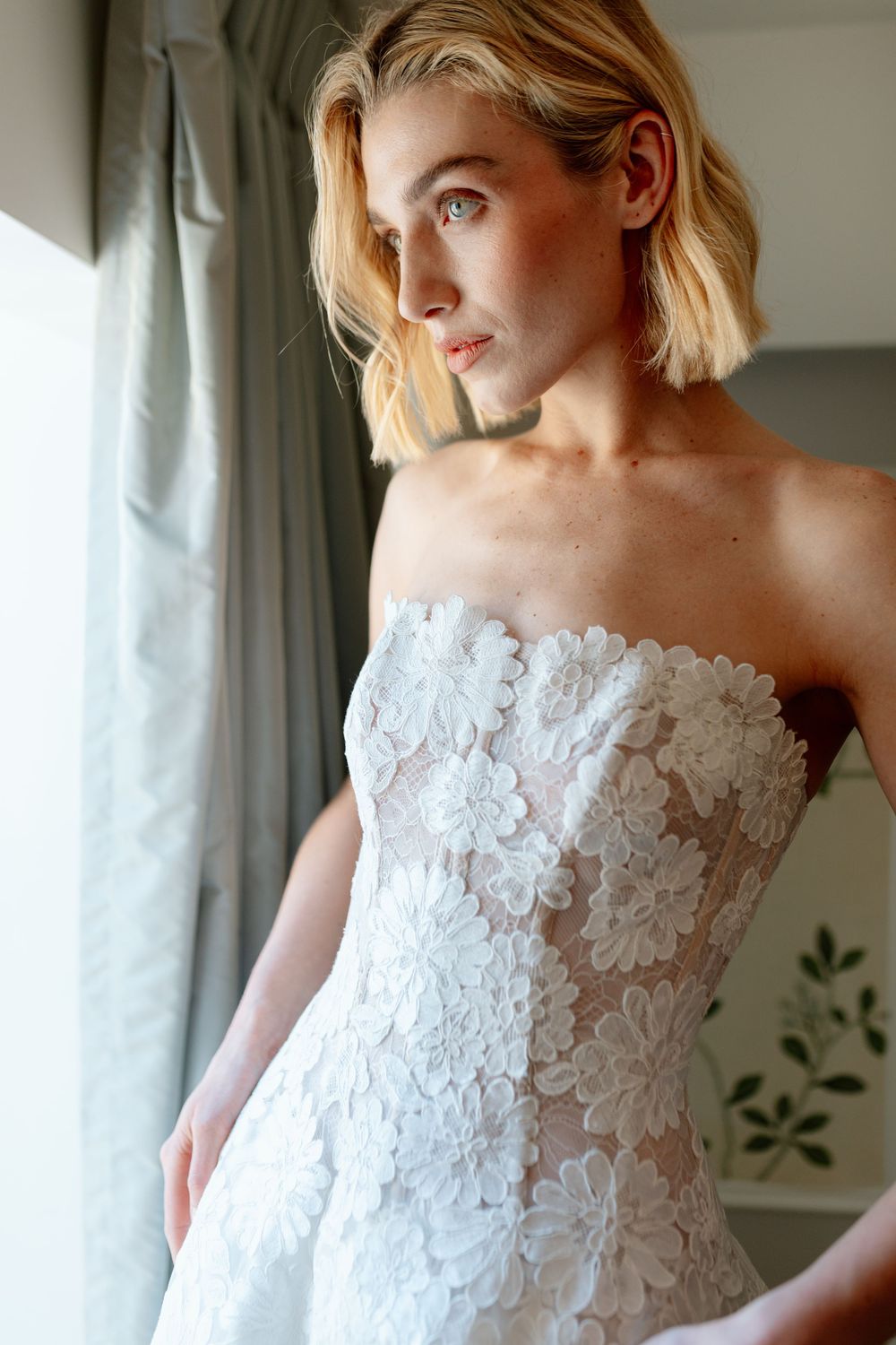 Blonde woman in a strapless white floral lace wedding gown posing near a window with soft natural light.