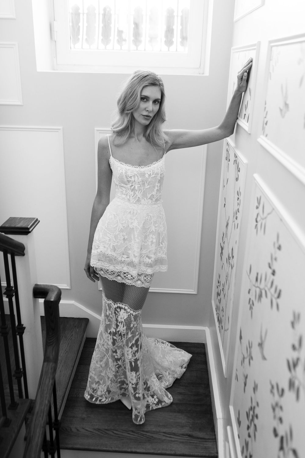 Blonde woman in white lace bridal mini dress with sheer floral train posing on staircase, black and white photo.